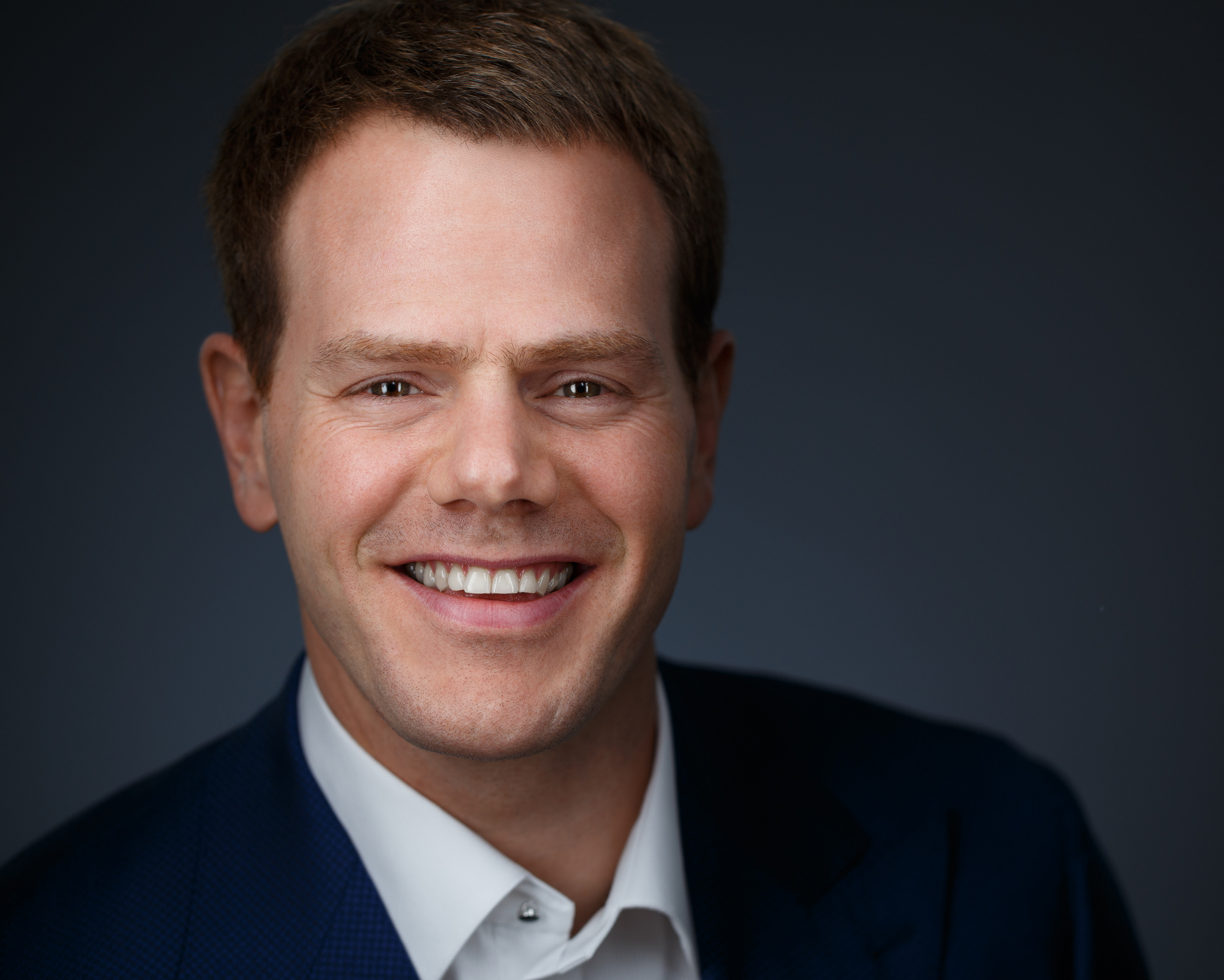 Бизнес-хедшот мужчины с улыбкой на тёмном фоне.