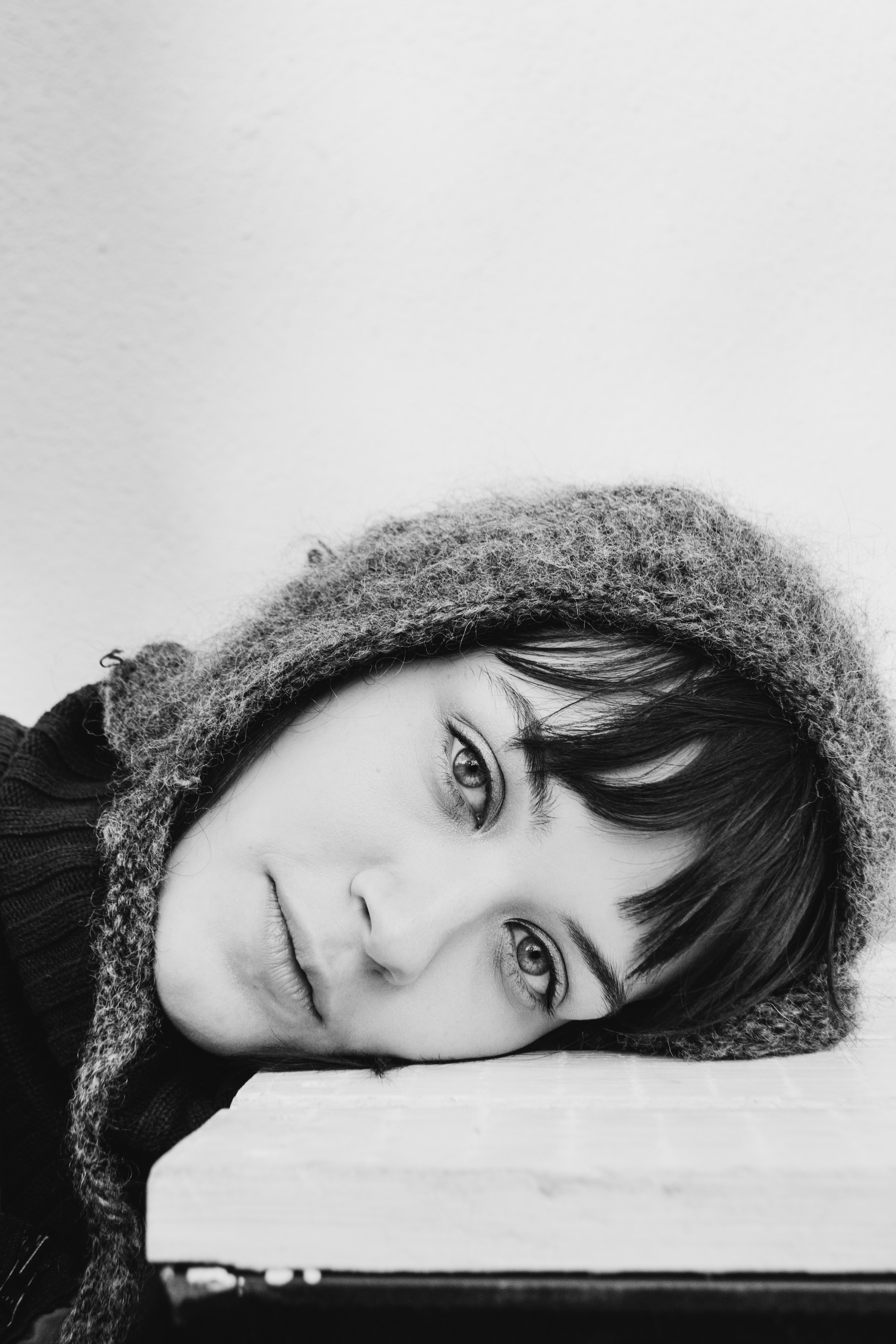 Зимний черно-белый крупный портрет девушки, чья голова лежит на столе. На модели вязаная шапка с завязками и челка. Она смотрит на нас