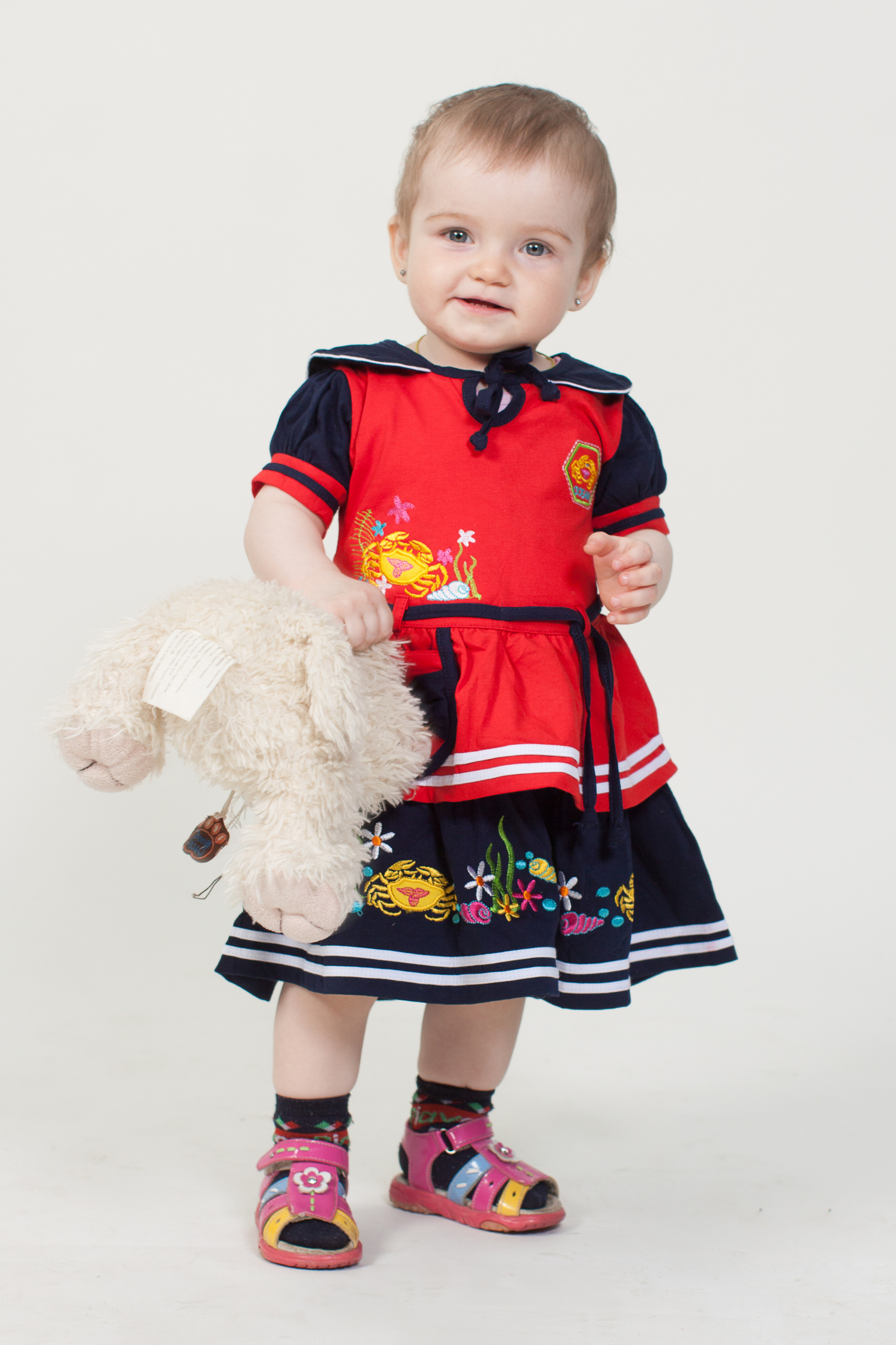 Девочка в красно-черном платье с мягкой игрушкой в руке на белом фоне.