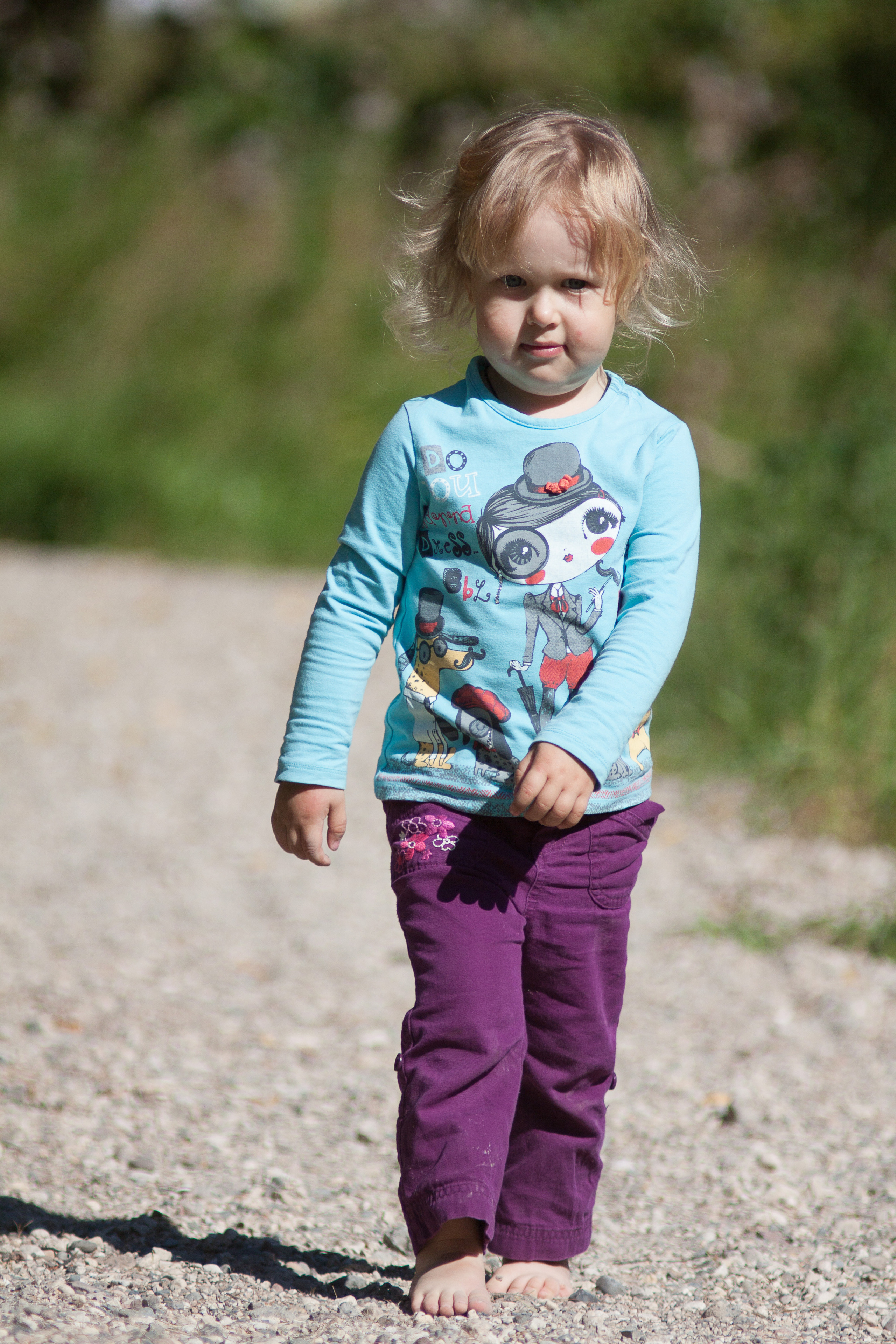 Девочка в бирюзовой кофте и малиновых брюках идет босиком по гравийной дорожке.