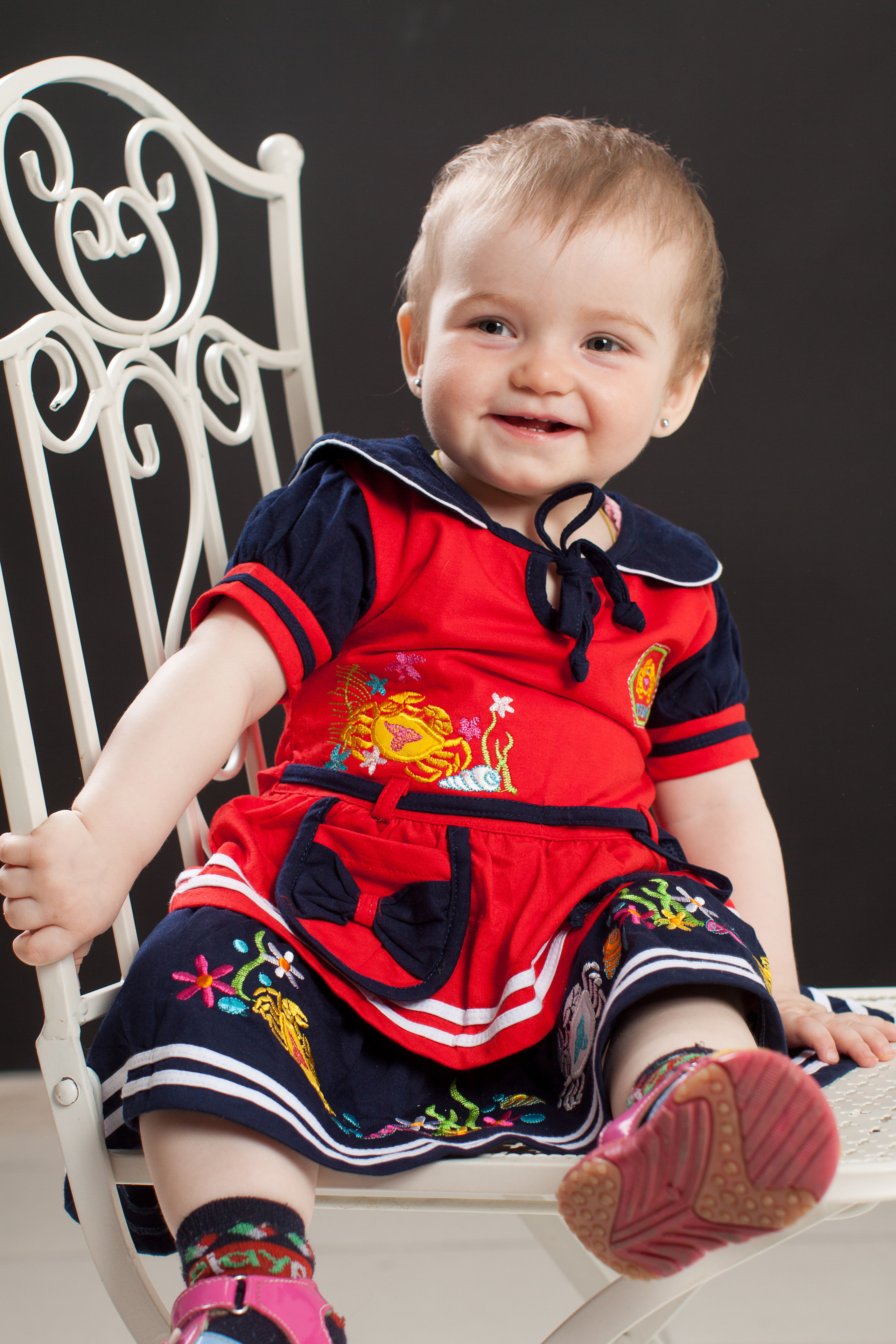 Девочка в красно-черном платье сидит на стуле. Задний фон черный.