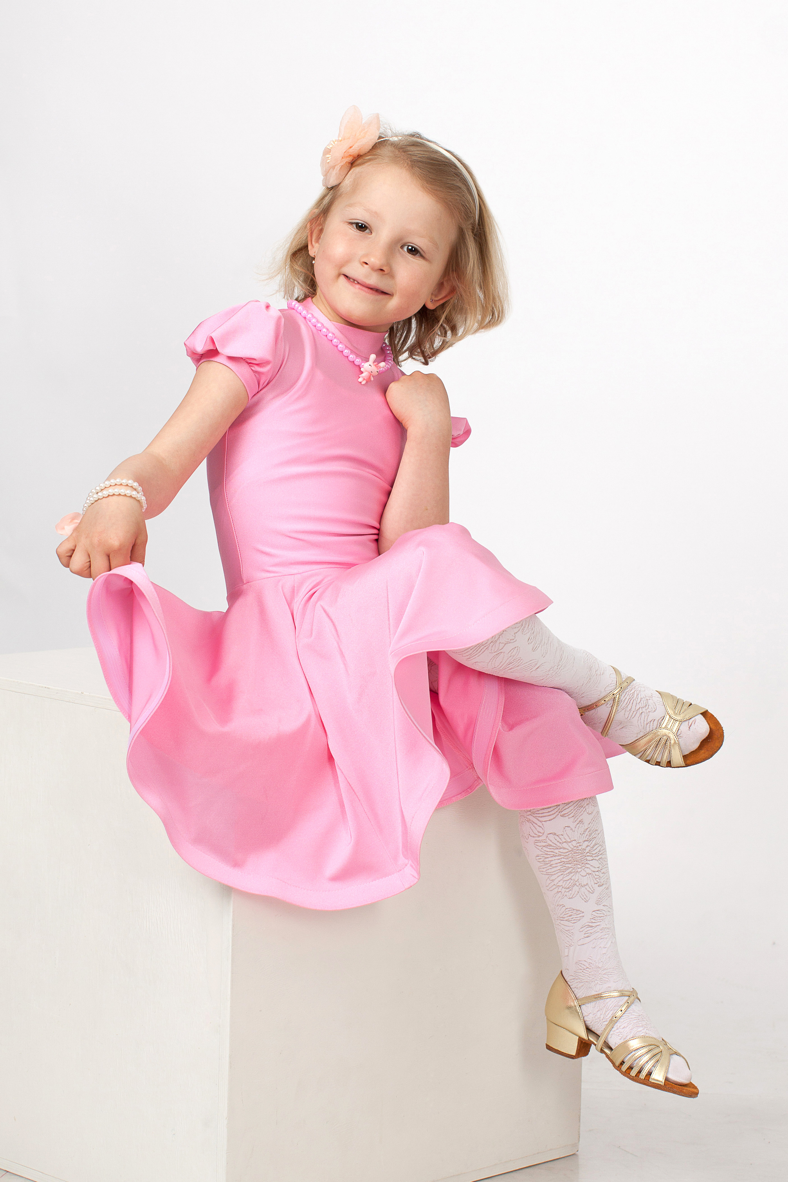 Девочка в розовом платье сидит на белом кубе на светлом фоне сзади.