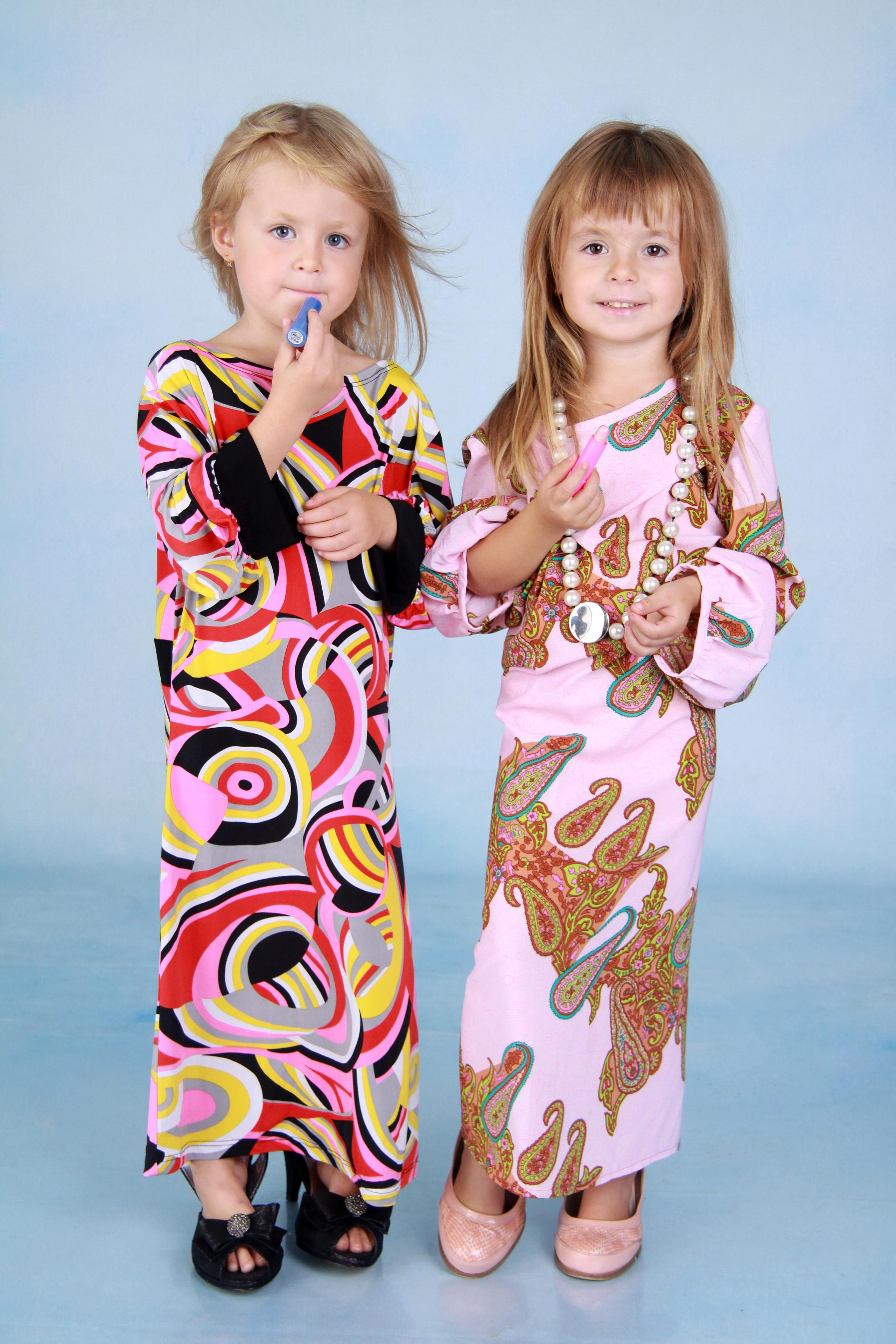 Две девочки 4-х лет стоя, одетые в маминых платьях с помадой в руках на фоне голубого студийного фона.