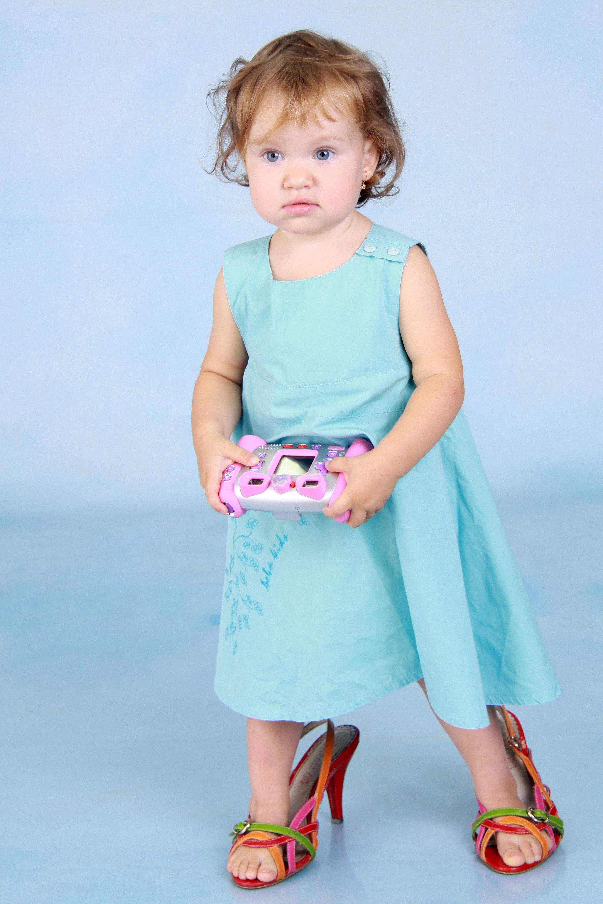 Девочка 2х лет стоит в маминых туфлях на фоне голубого студийного фона.