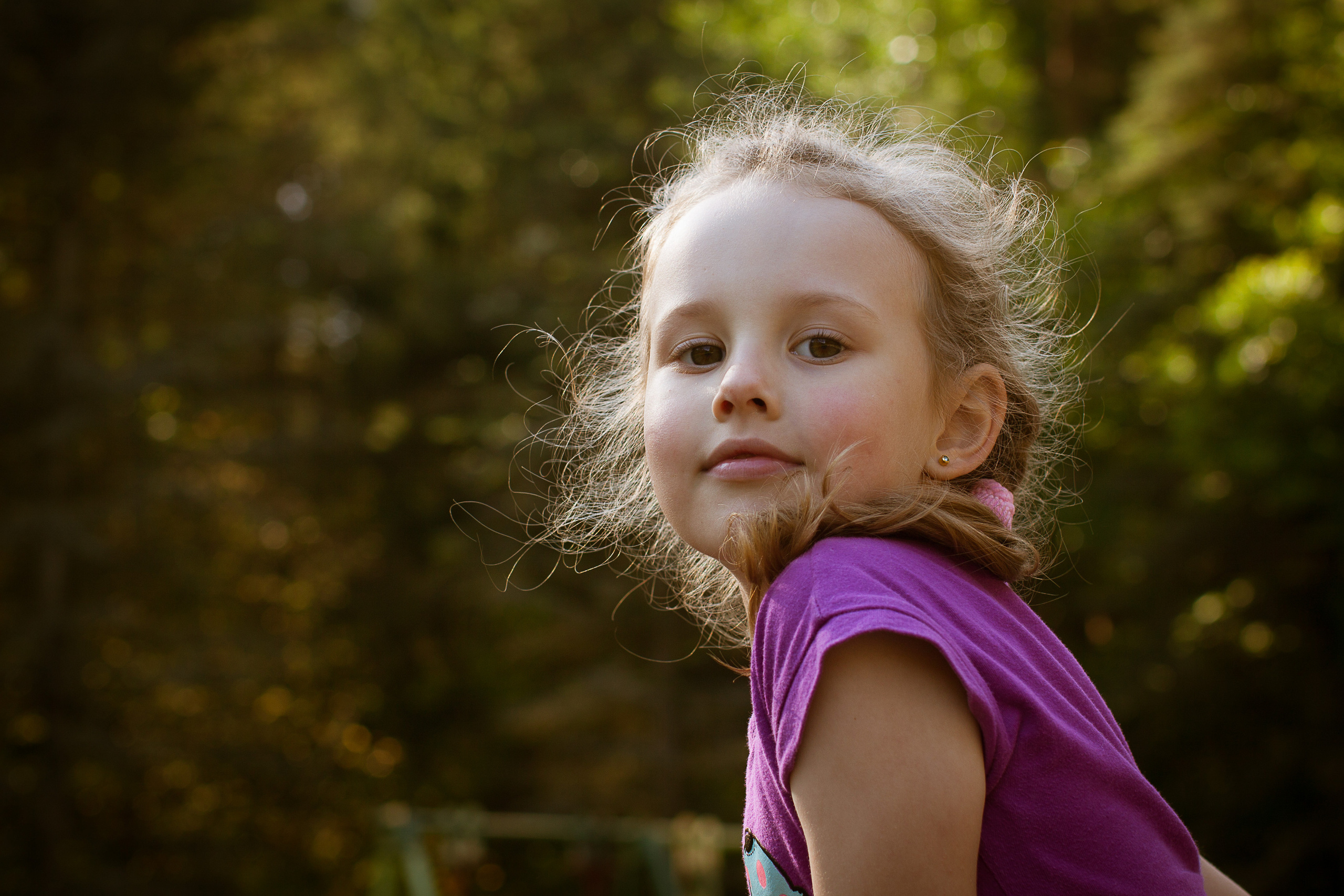 Девочка в малиновой футболке смотрит через плечо на фоне деревьевд