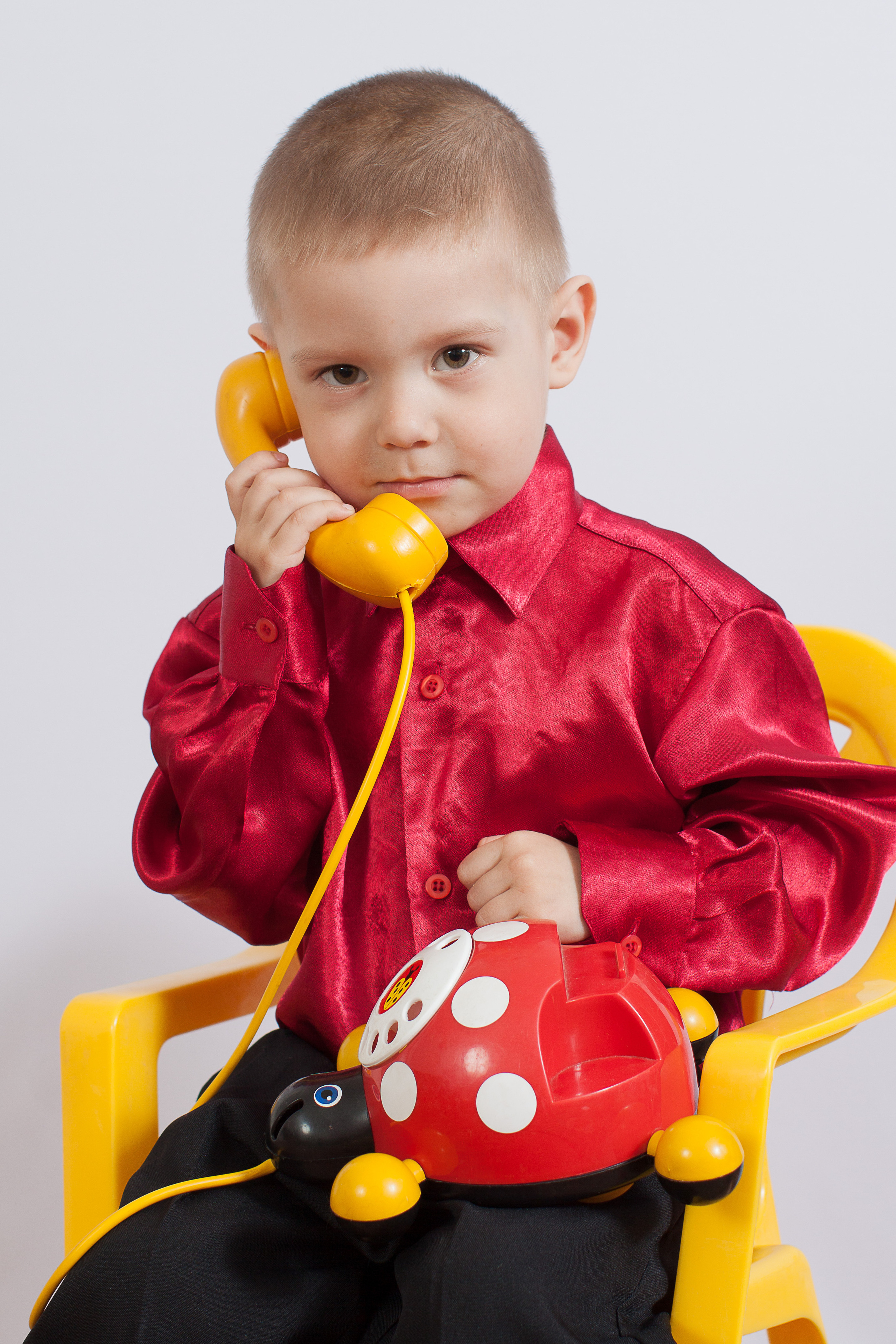 Мальчик сидит на желтом стуле  в красной рубашке с желтым телефоном в руке.