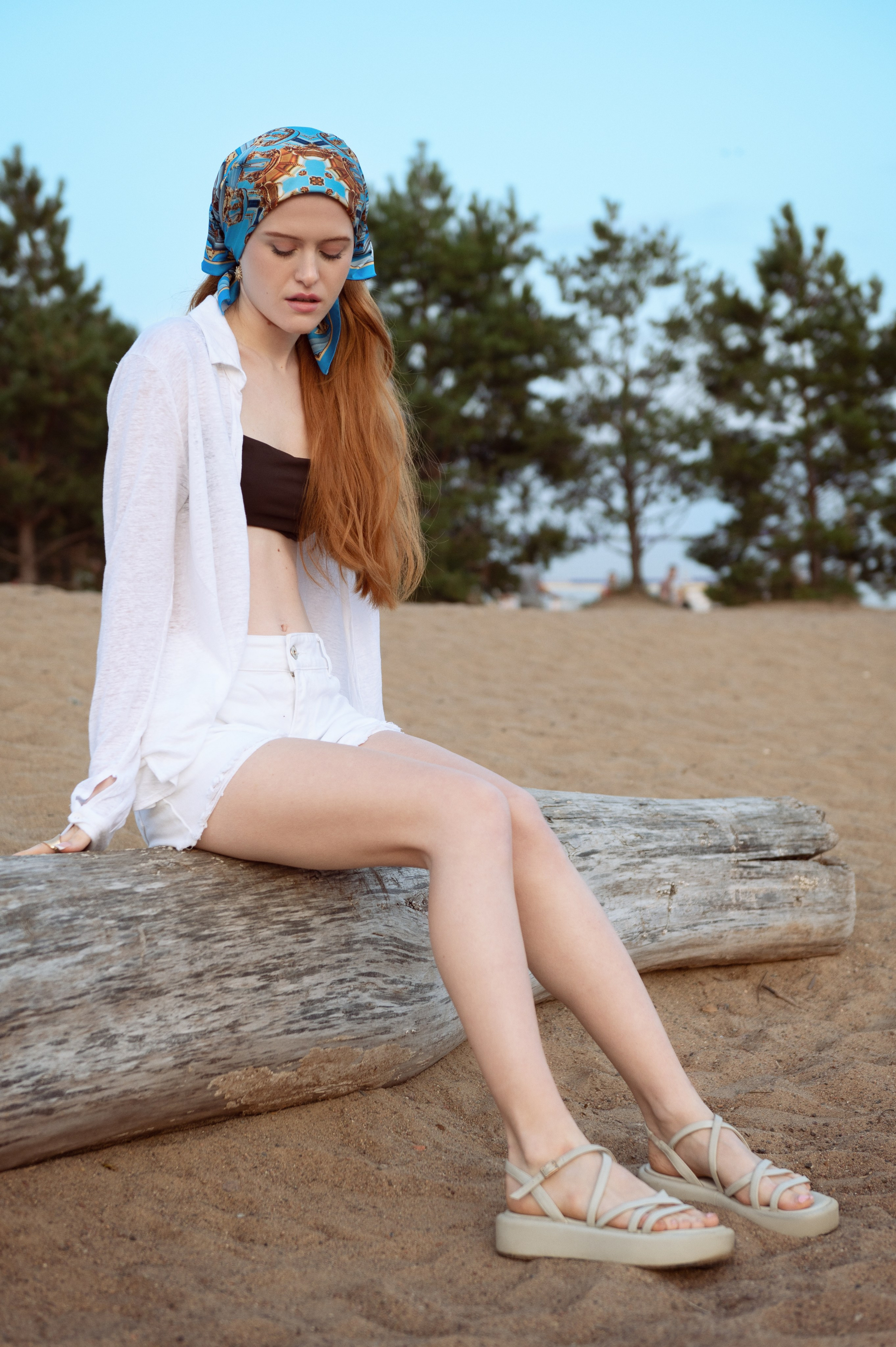 Красивая рыжеволосая девушка в белой рубашке и белых шортах сидит на песчаном пляже