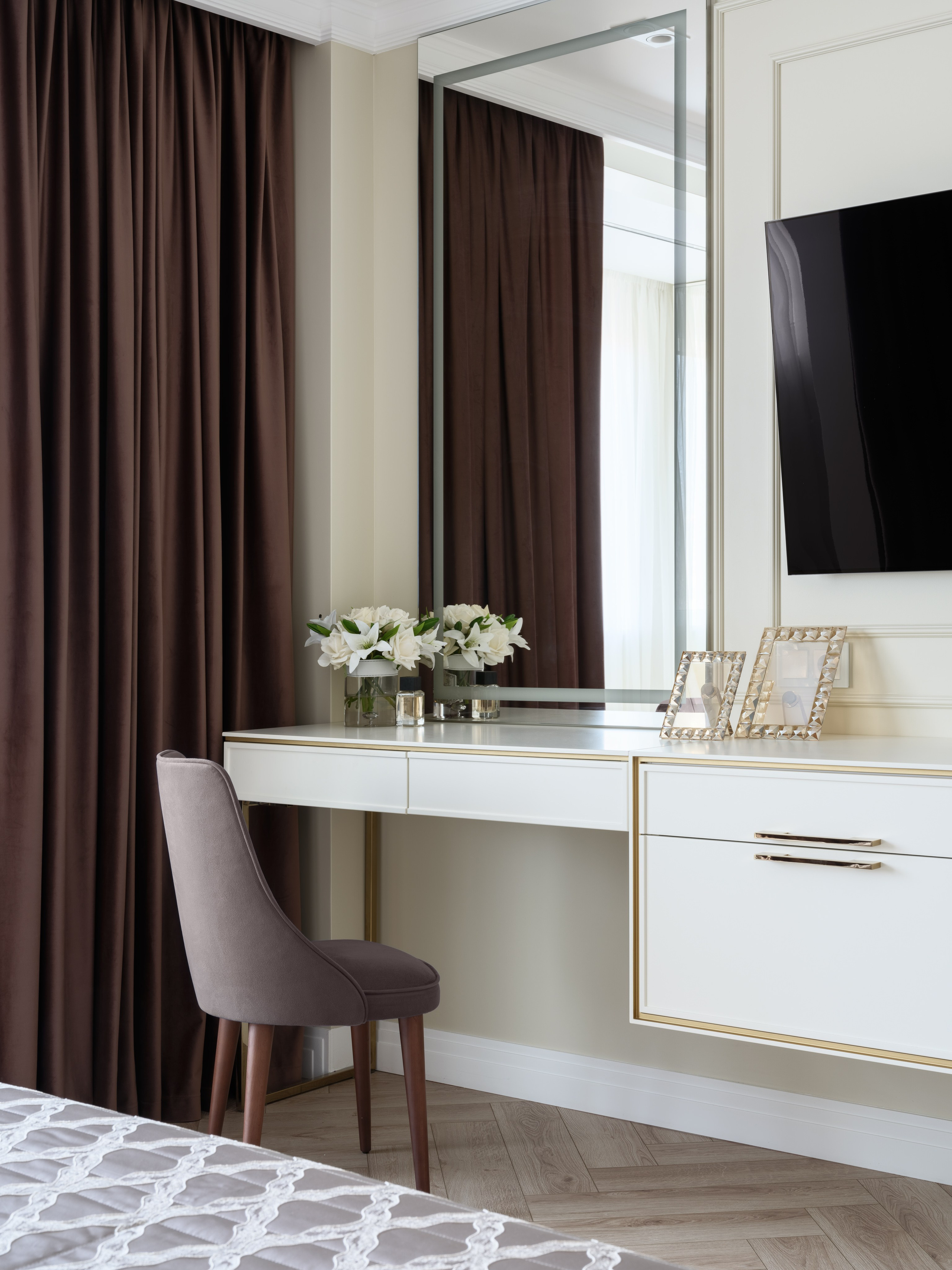 Фото современного дизайна интерьера спальни с туалетным столиком и зеркалом во всю стену, белыми фасадами с золотой окантовкой и серым мягким стулом. На столике букет белых цветов и фоторамки, создающие уют и гармонию. Интерьер разработан дизайнером Евгенией Ермошиной и сфотографирован Сергеем Денисенко.