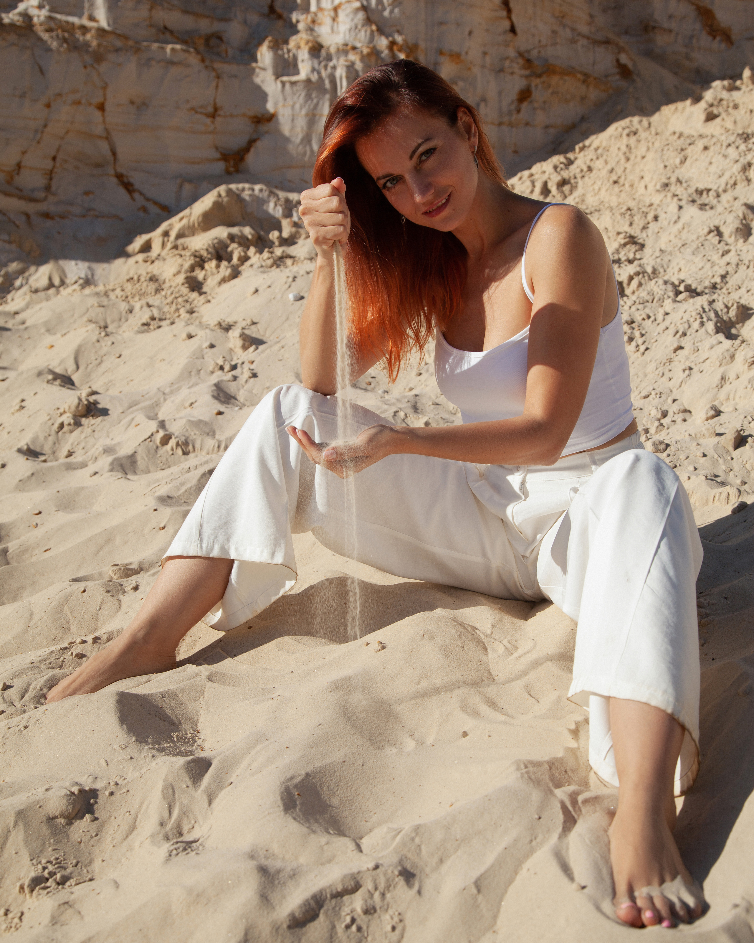 Рыжая девушка сидит на песке в пустыне, на ней одеты белые брюки