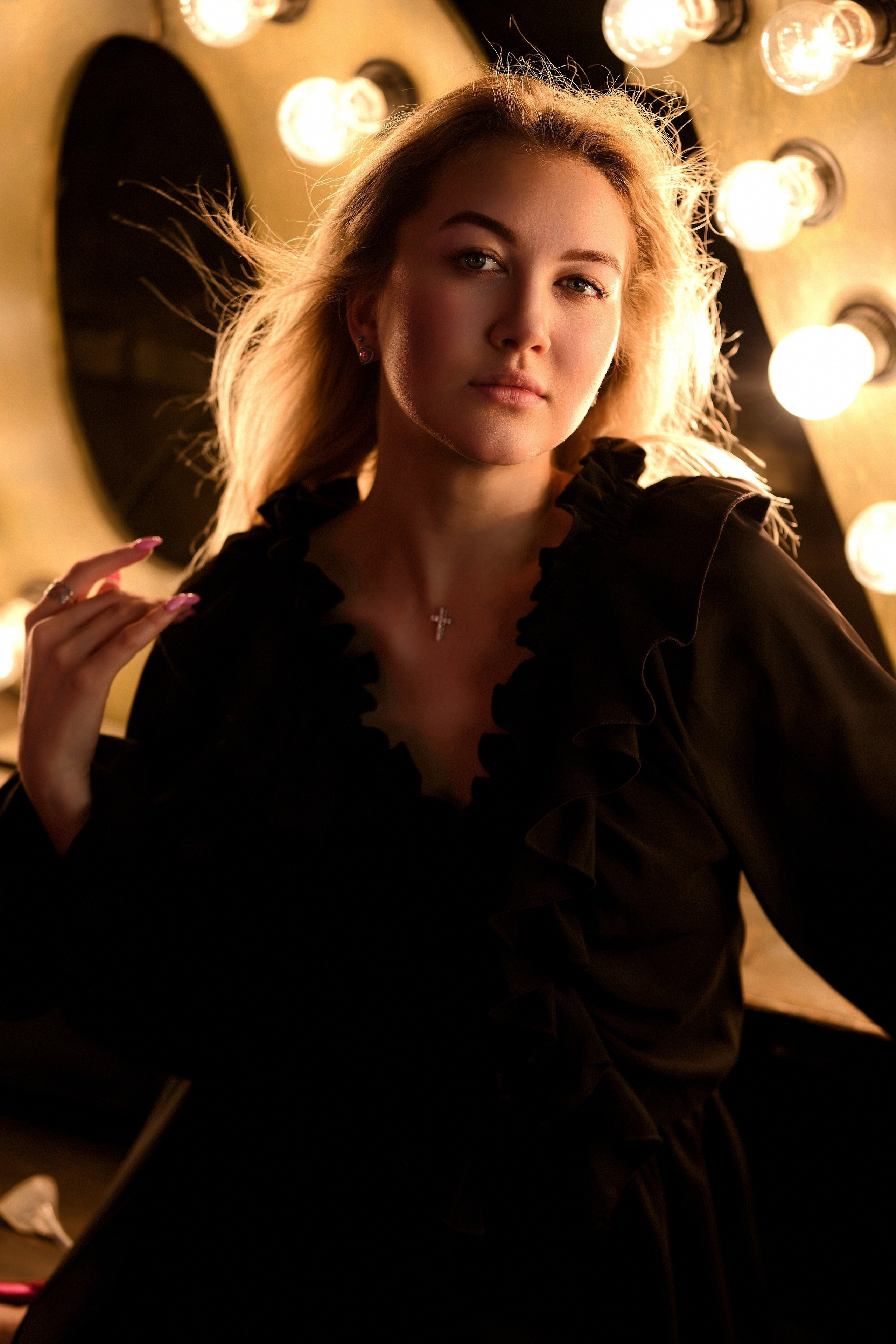 Портрет блондинки в черном платье с оборками на фоне гримерного зеркала с лампочками, контровой свет, развевающиеся волосы, крестик на шее