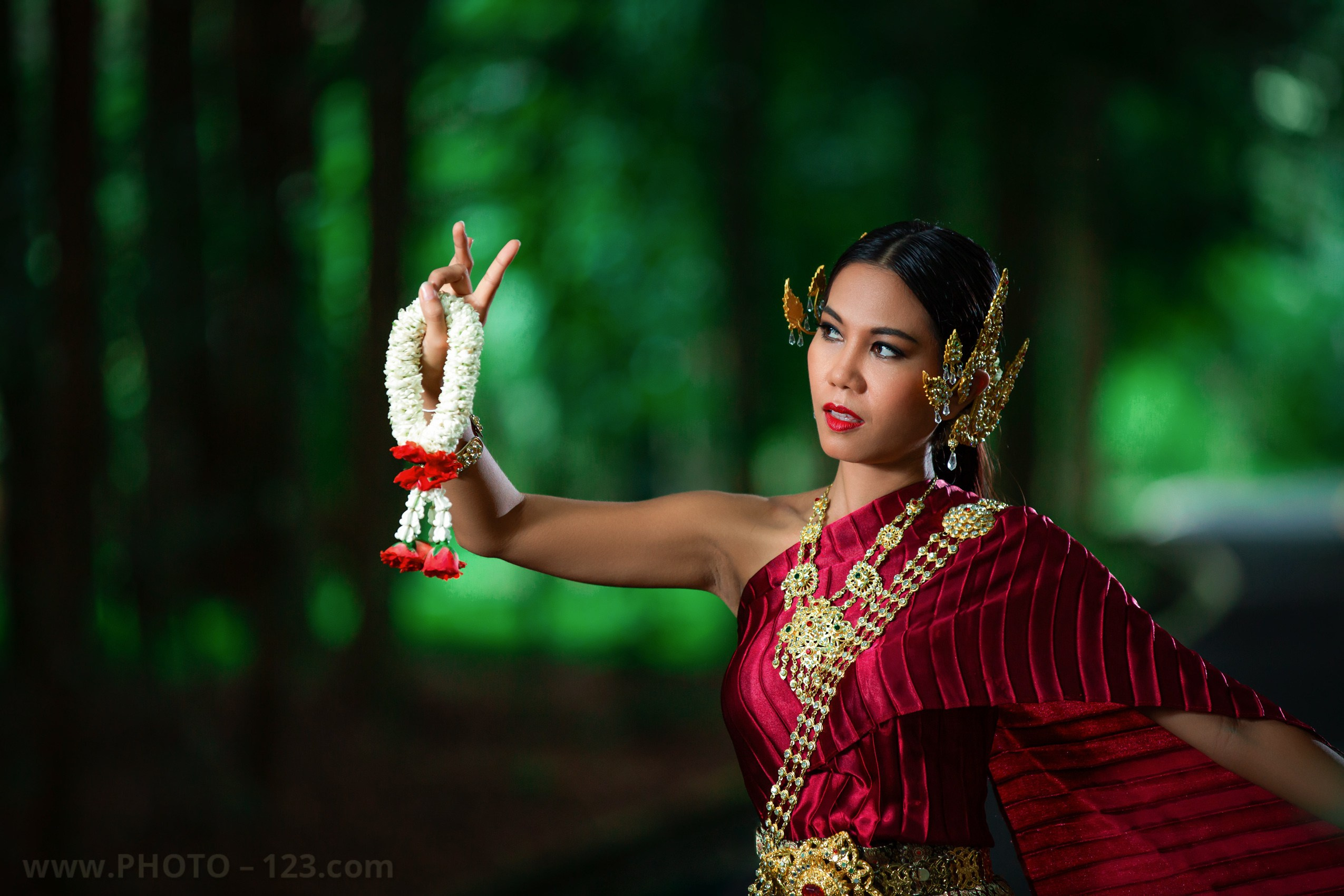 Portrait of a Thai dancer in traditional maroon and gold costume performing classical gesture with floral garland; surrounded by lush greenery, ornate shrine, and sunset light; editorial-style image blending cultural heritage and graceful movement, photographed in Phu Quoc