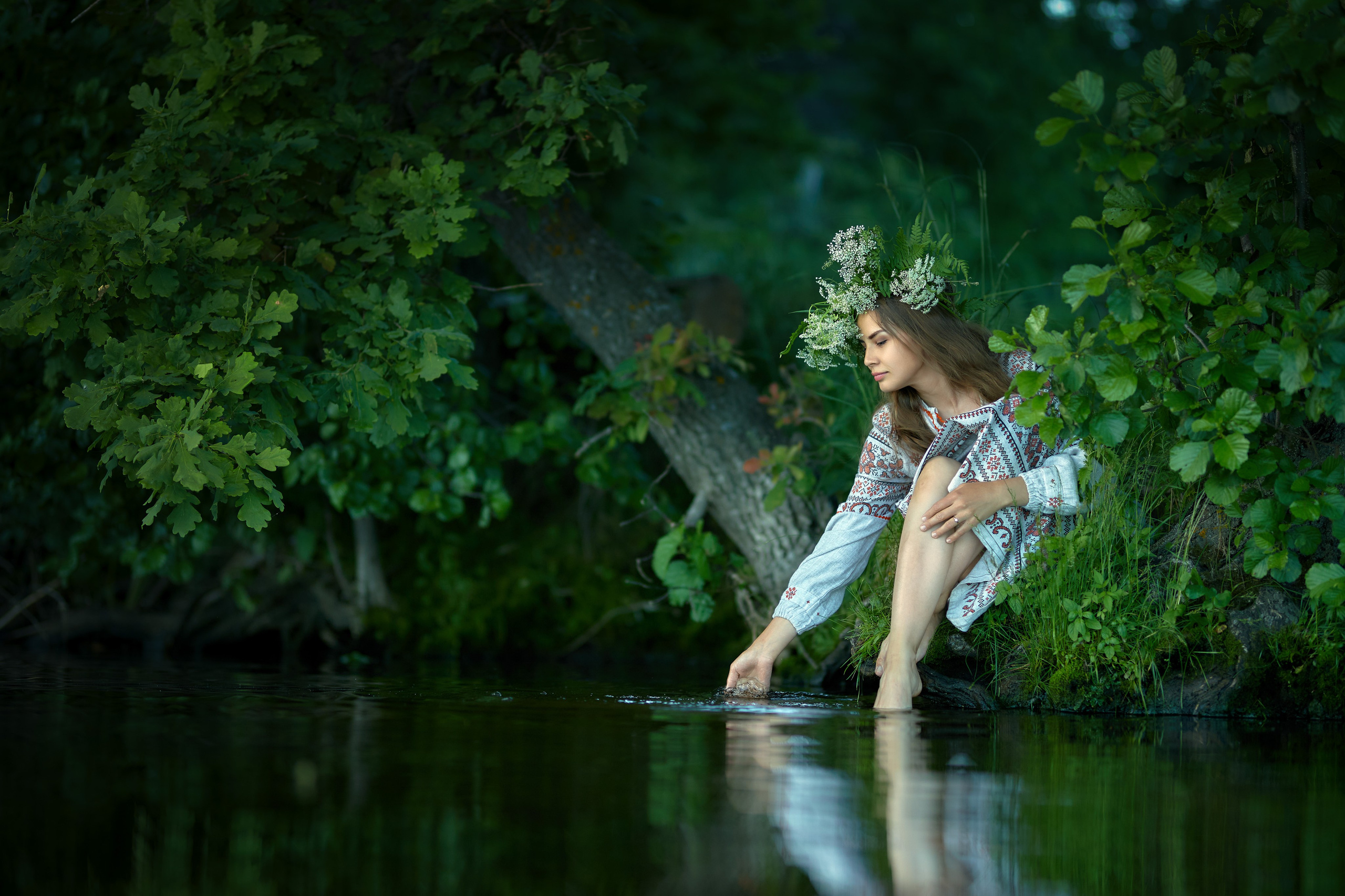 Девушка в национальном костюме и венком на голове сидит у воды