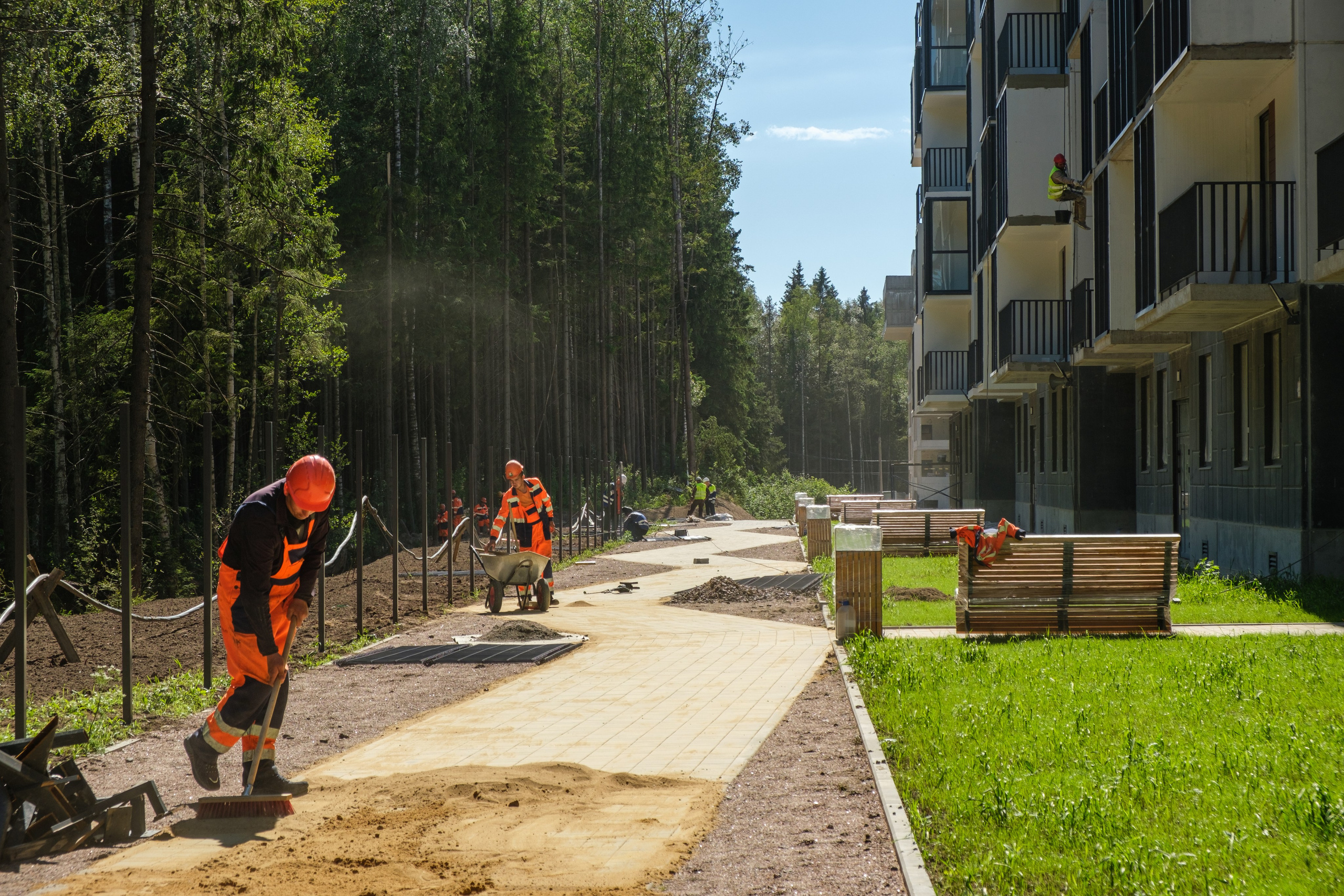 На фотографии изображены рабочие в спецодежде, занимающиеся укладкой тротуара. Один из рабочих использует лопату для распределения материала. Рядом с ними видны инструменты и материалы, необходимые для выполнения работ.