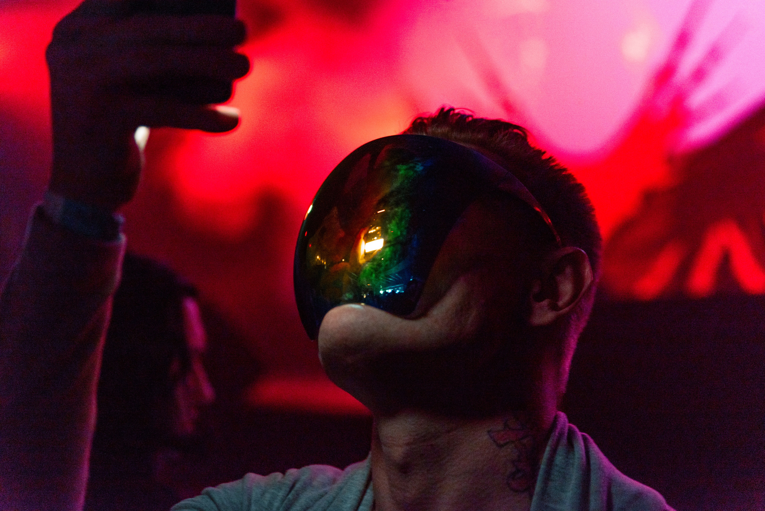 Концерт, лицо мужчины закрытое зеркальной маской, в руке напротив лица держит телефон, на заднем плане сцена освещённая красным светом