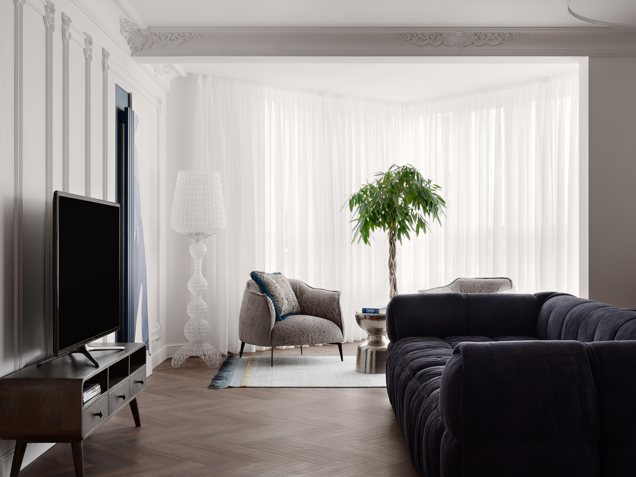 Современный дизайн интерьера гостиной с уютной атмосферой: стильный фотообзор зоны отдыха с мягким тёмным диваном, телевизором на тумбе и угловой зоной с креслами у окна. Автор проекта — дизайнер Антон Логинов, стилист Алёна Мощенская, фотограф Сергей Денисенко.