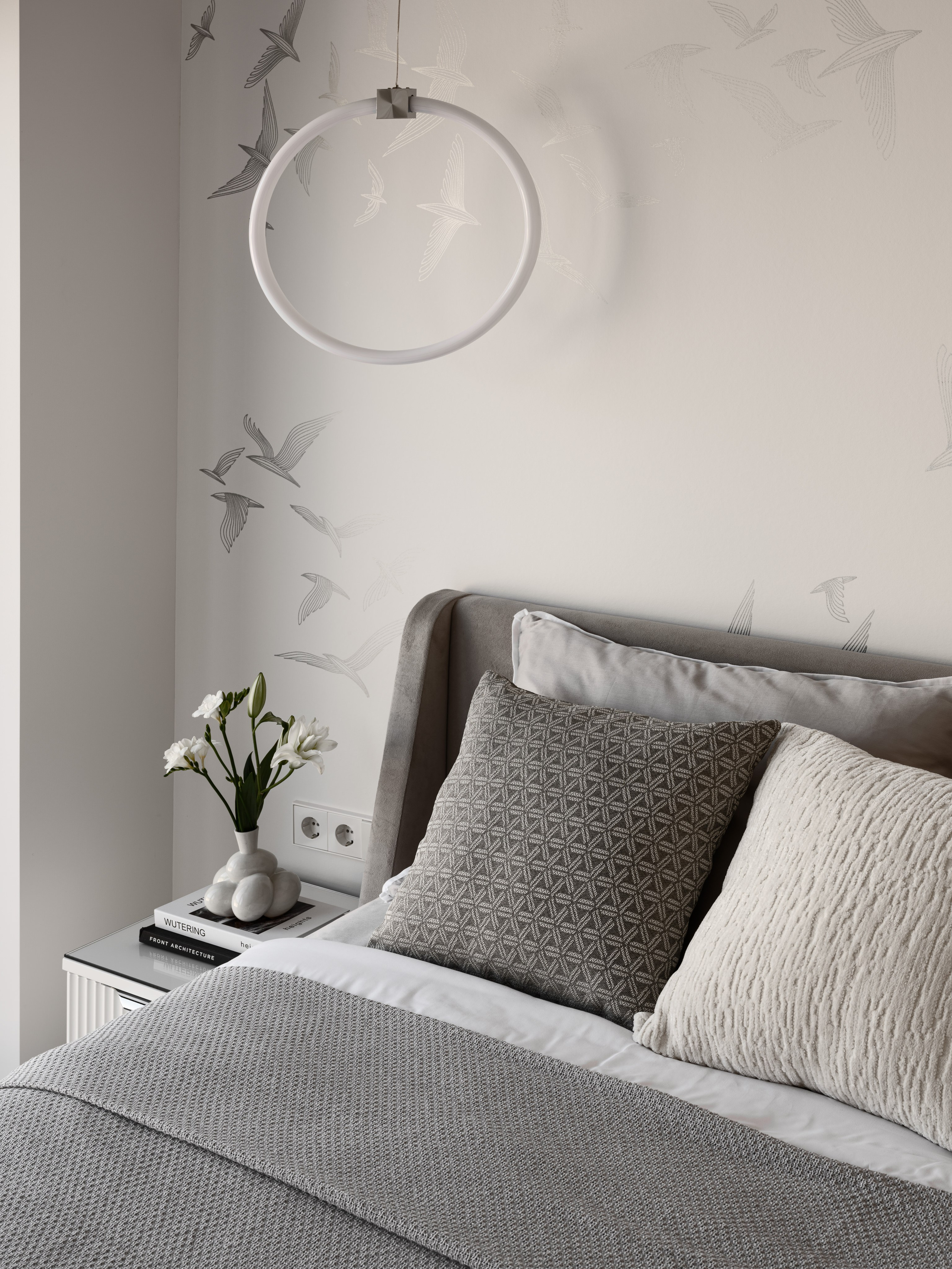 Элегантный интерьер спальни в светлых тонах с мягким изголовьем кровати, графичными подушками, минималистичной люстрой-кольцом и цветами в керамической вазе на прикроватной тумбе. Дизайнер Алёна Боярская, стилист Наталья Якушева, фотограф Сергей Денисенко.