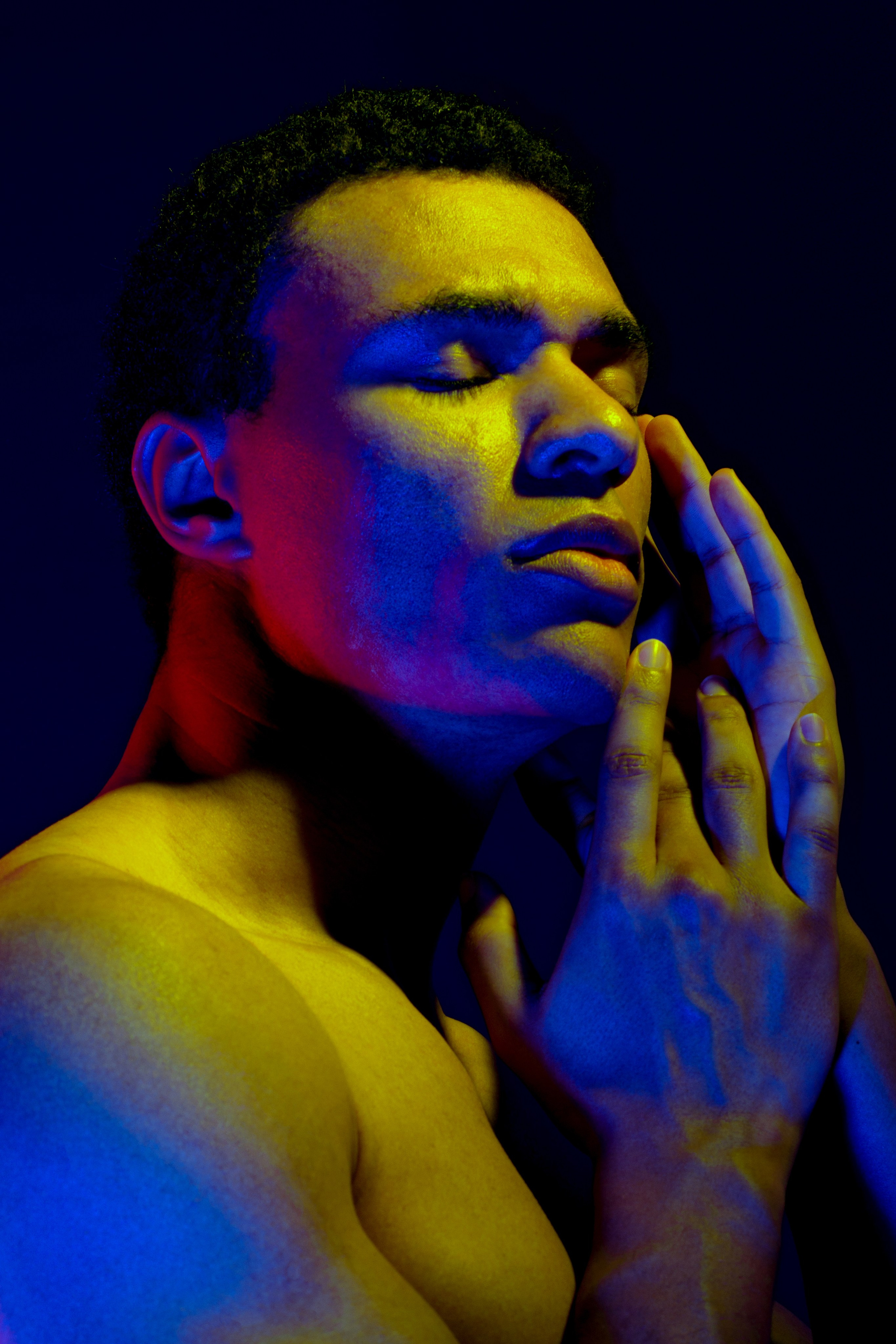 чернокожий обнаженный мужчина на черном фоне в цветном свете с руками у лица 