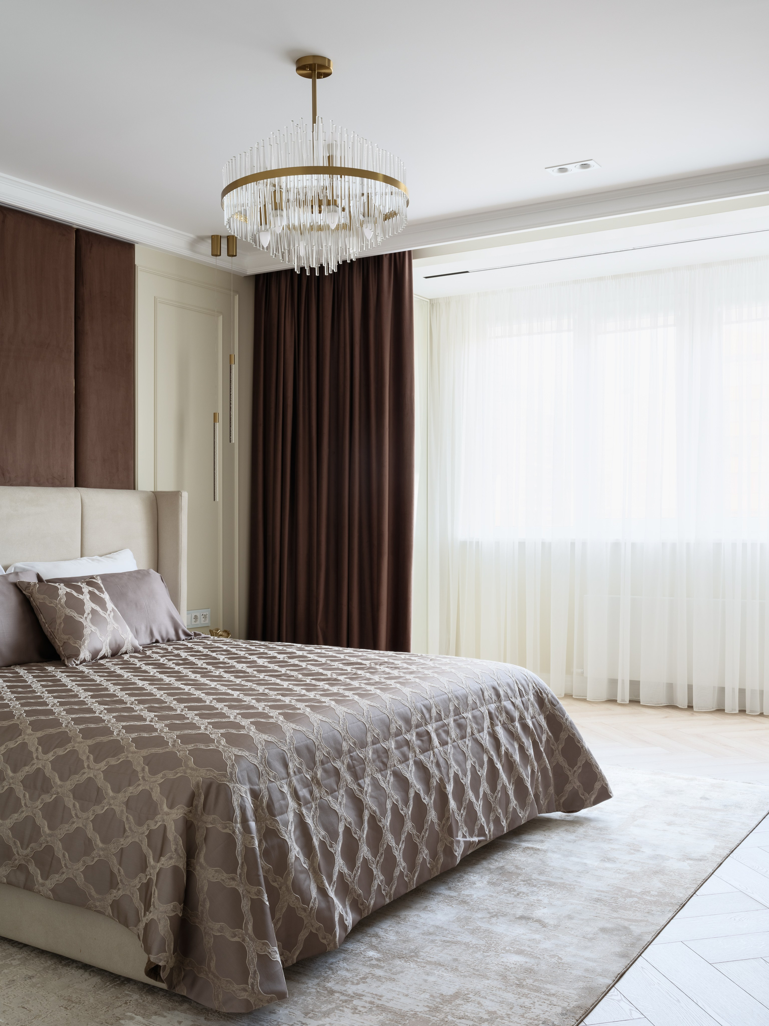 Просторная спальня в современном стиле с большим окном, легкими белыми и коричневыми шторами, светлым ковром и элегантной люстрой с хрустальными подвесами. Фото подчеркивает изысканный дизайн интерьера, созданный Евгенией Ермошиной и сфотографированный Сергеем Денисенко.