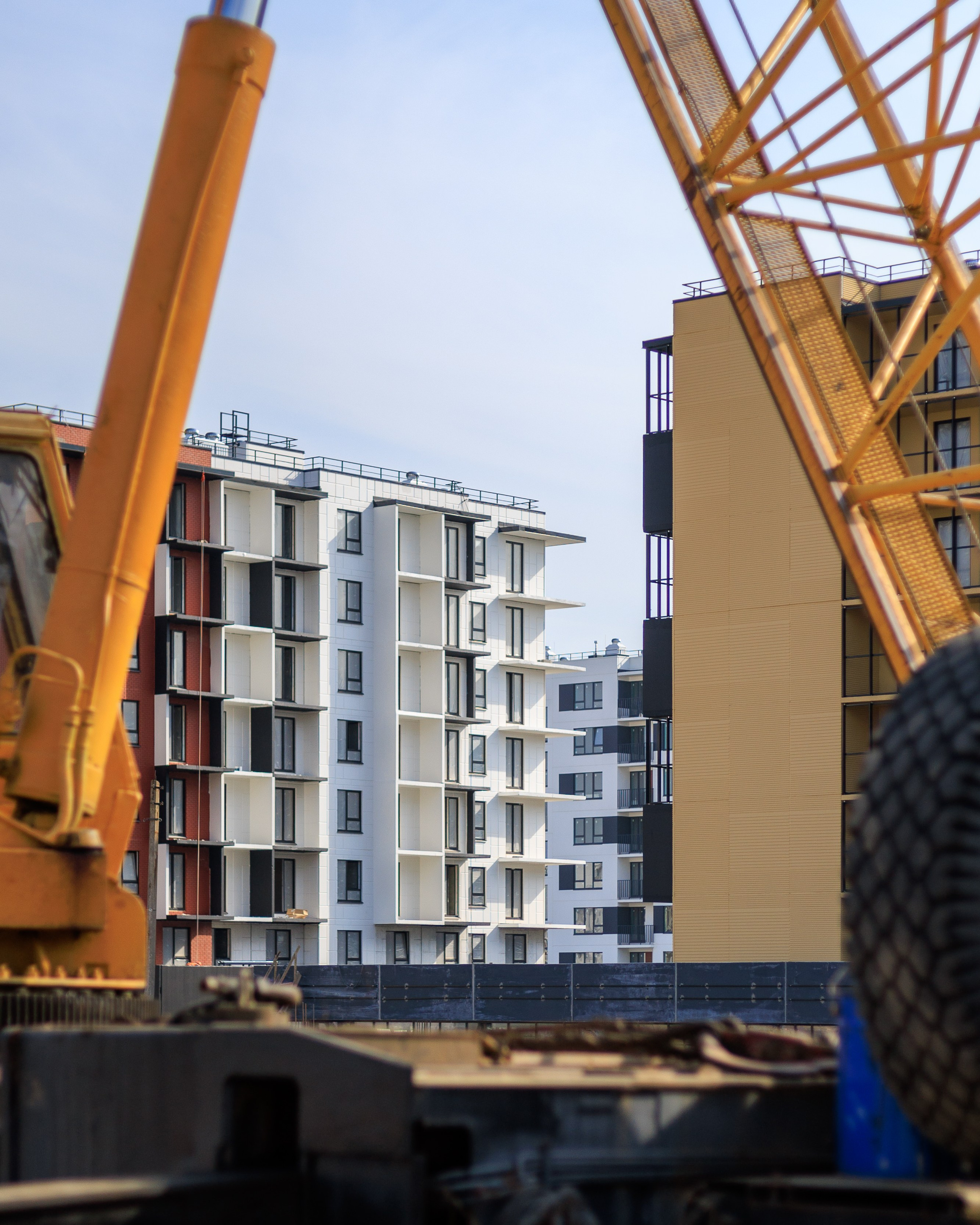 На изображении представлена строительная площадка, где ведутся работы по возведению многоэтажного жилого комплекса. Видны строительные краны и частично построенные здания.