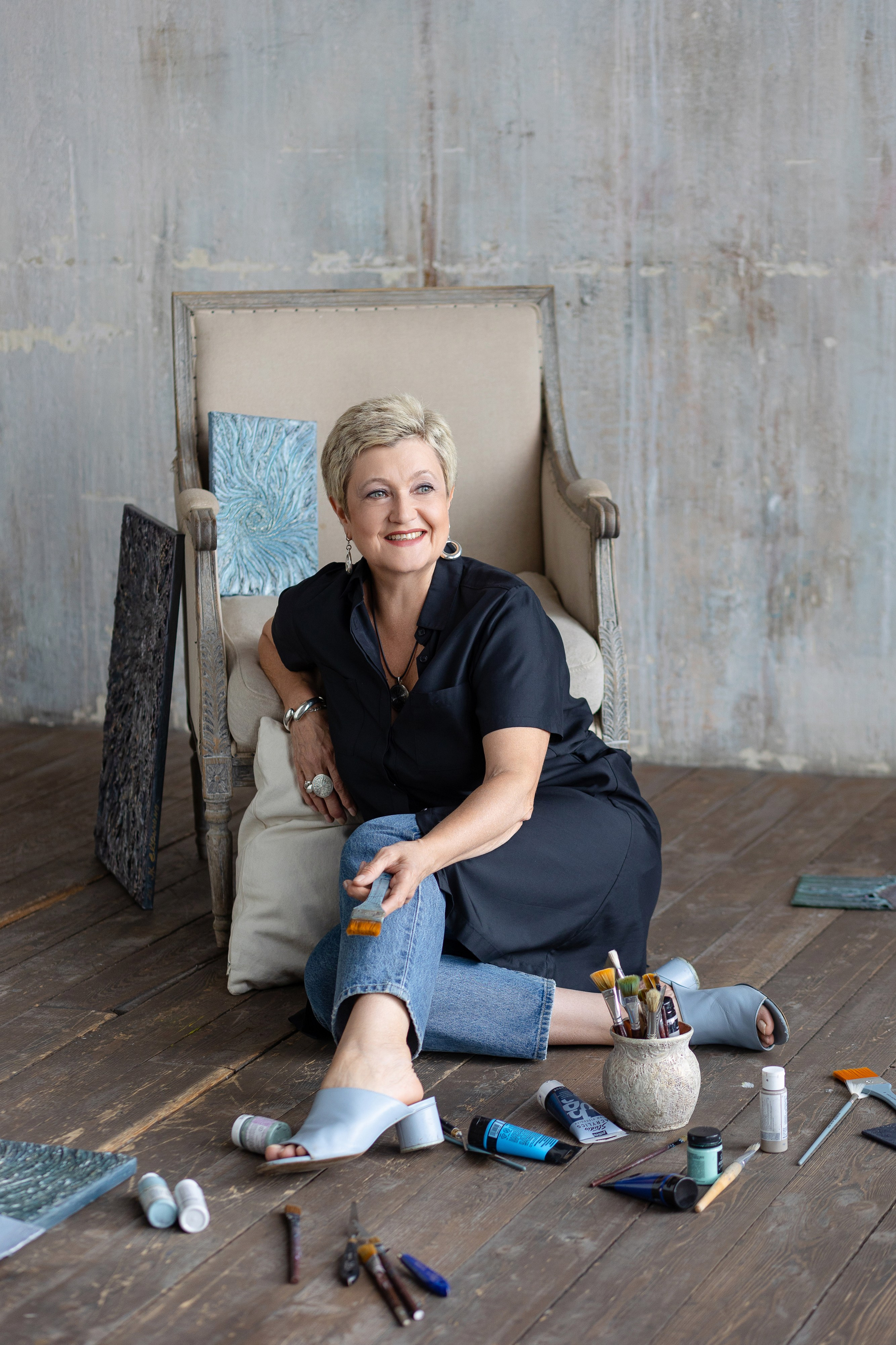 Художница сидит на полу возле кресла, перед ней кисти и тюбики с краской.