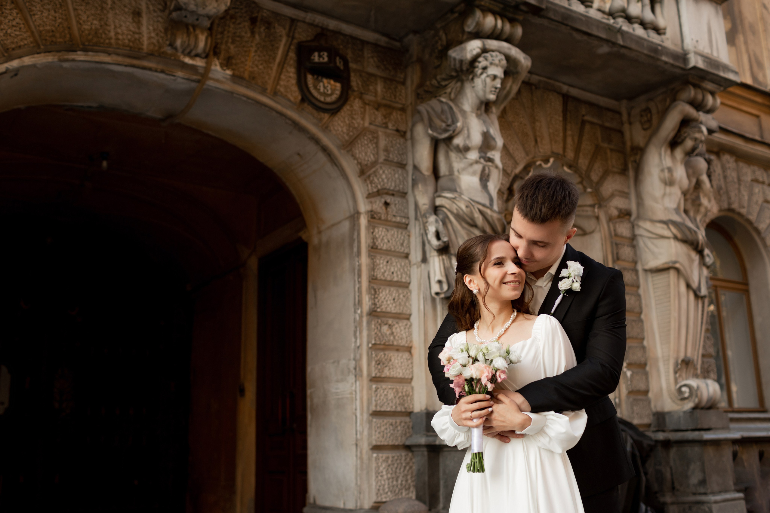 Живая фотография молодоженов: жених обнимает невесту на фоне исторического здания Петербурга.