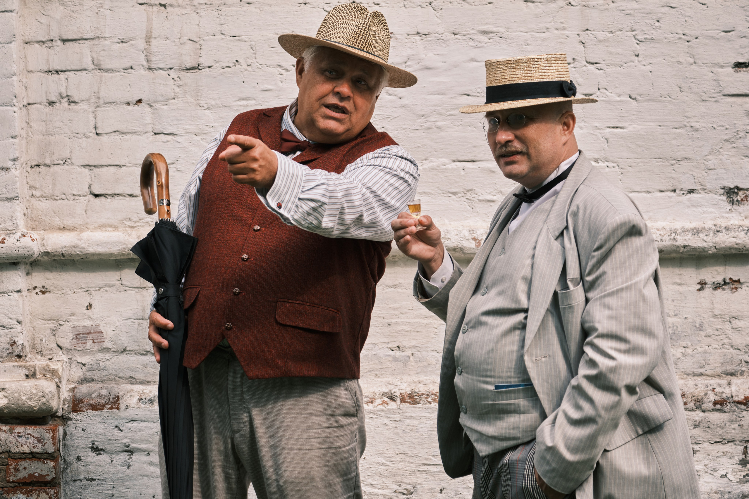 Двое мужчин в шляпах обсуждают что то показывая пальцем,  у одного мужчины рюмка с коньяком