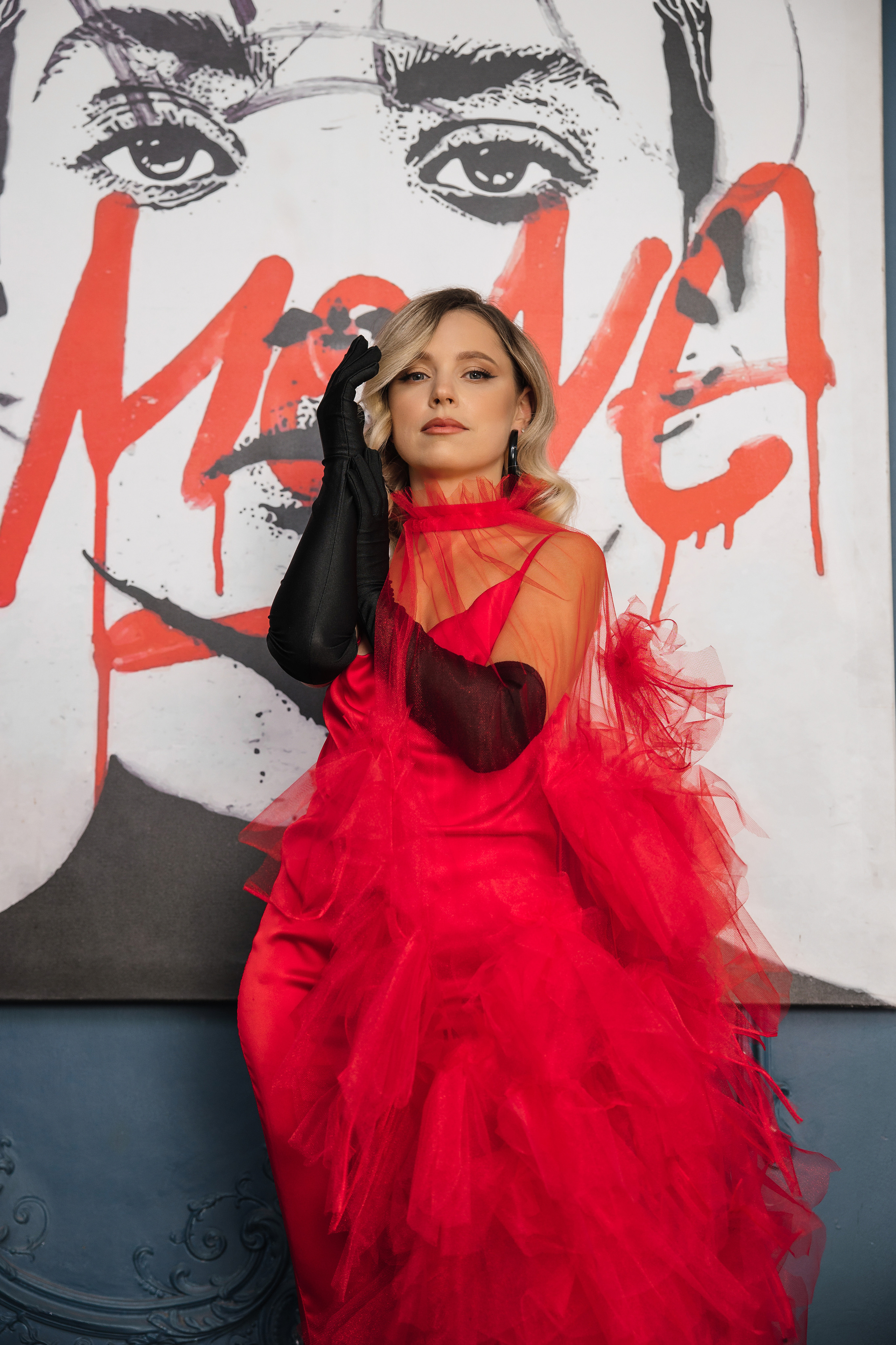 Привлекательная блондинка в ярком красном платье и красных перчатках на фоне картины с графити.