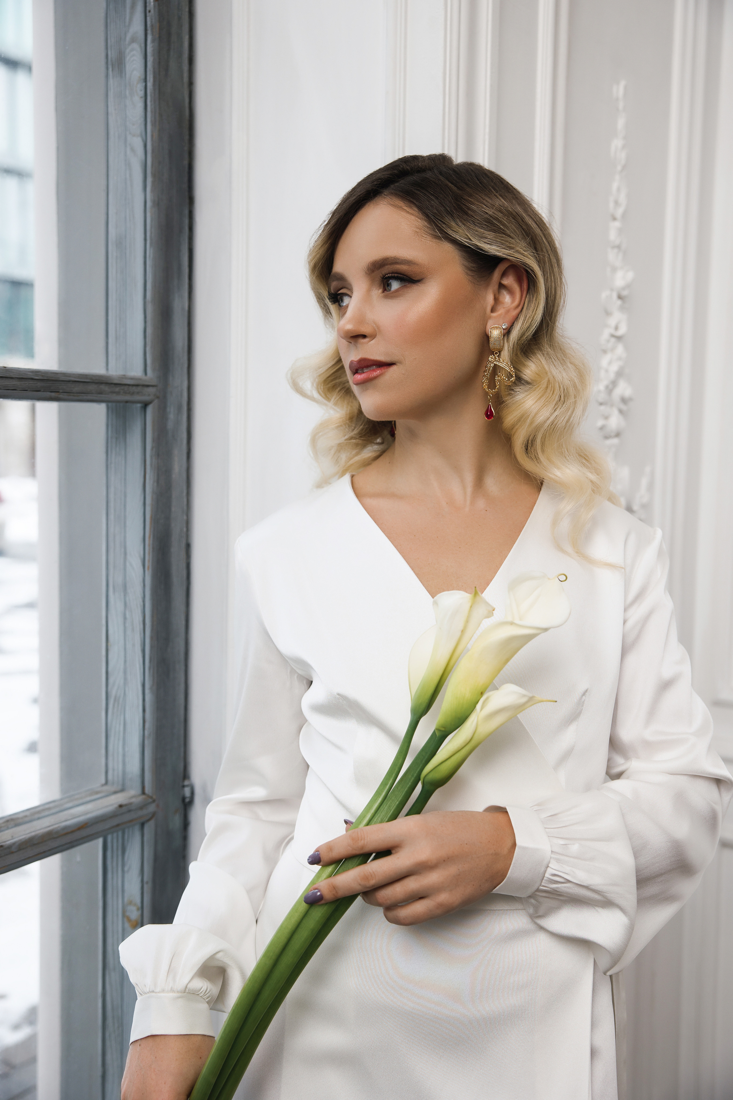 невеста в белом свадебном платье позирует у окна с букетом калл