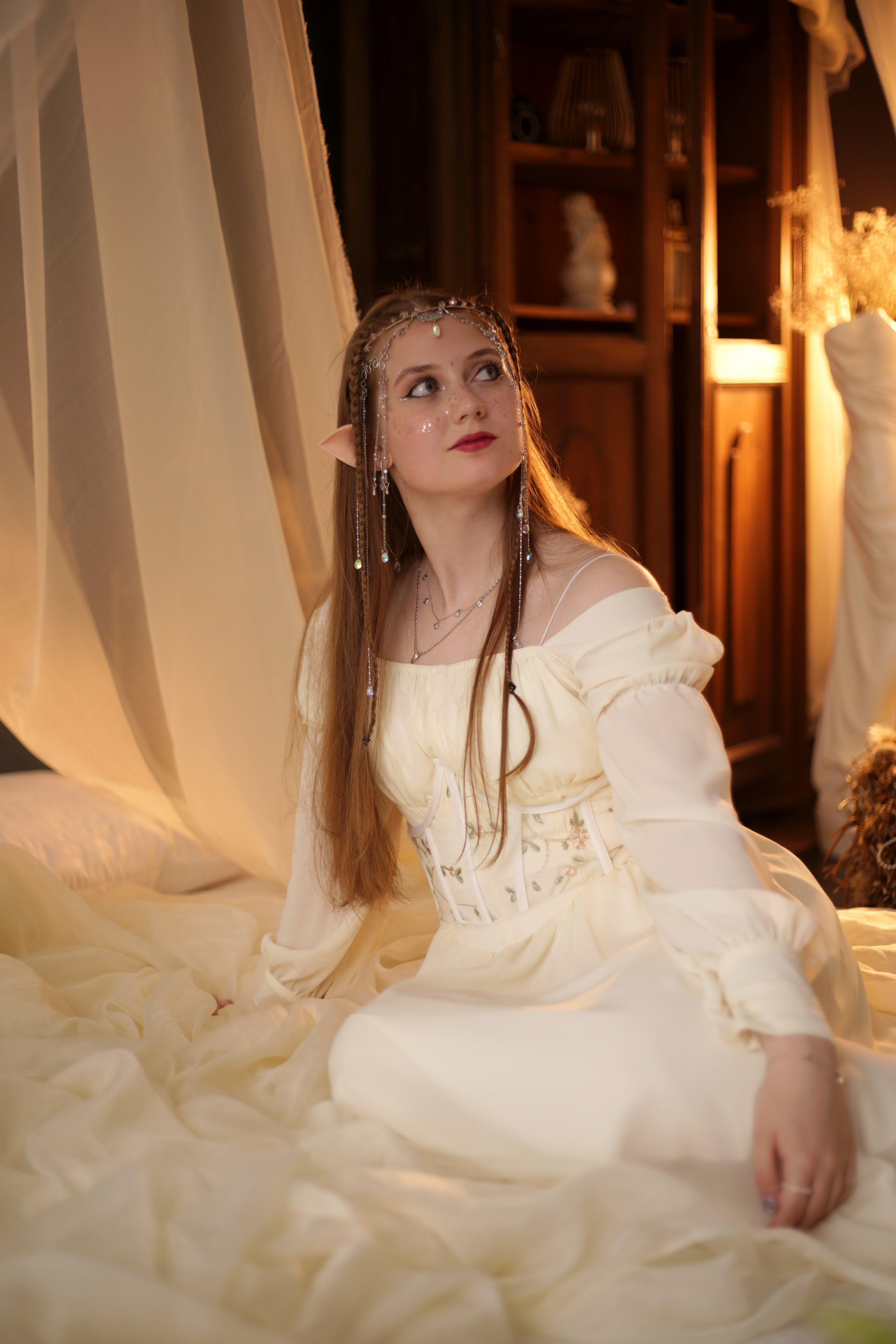 Эльфийская фотосессия в Перми: девушка в белом платье с ушками и головным убором, сидит на кровати под полупрозрачным балдахином