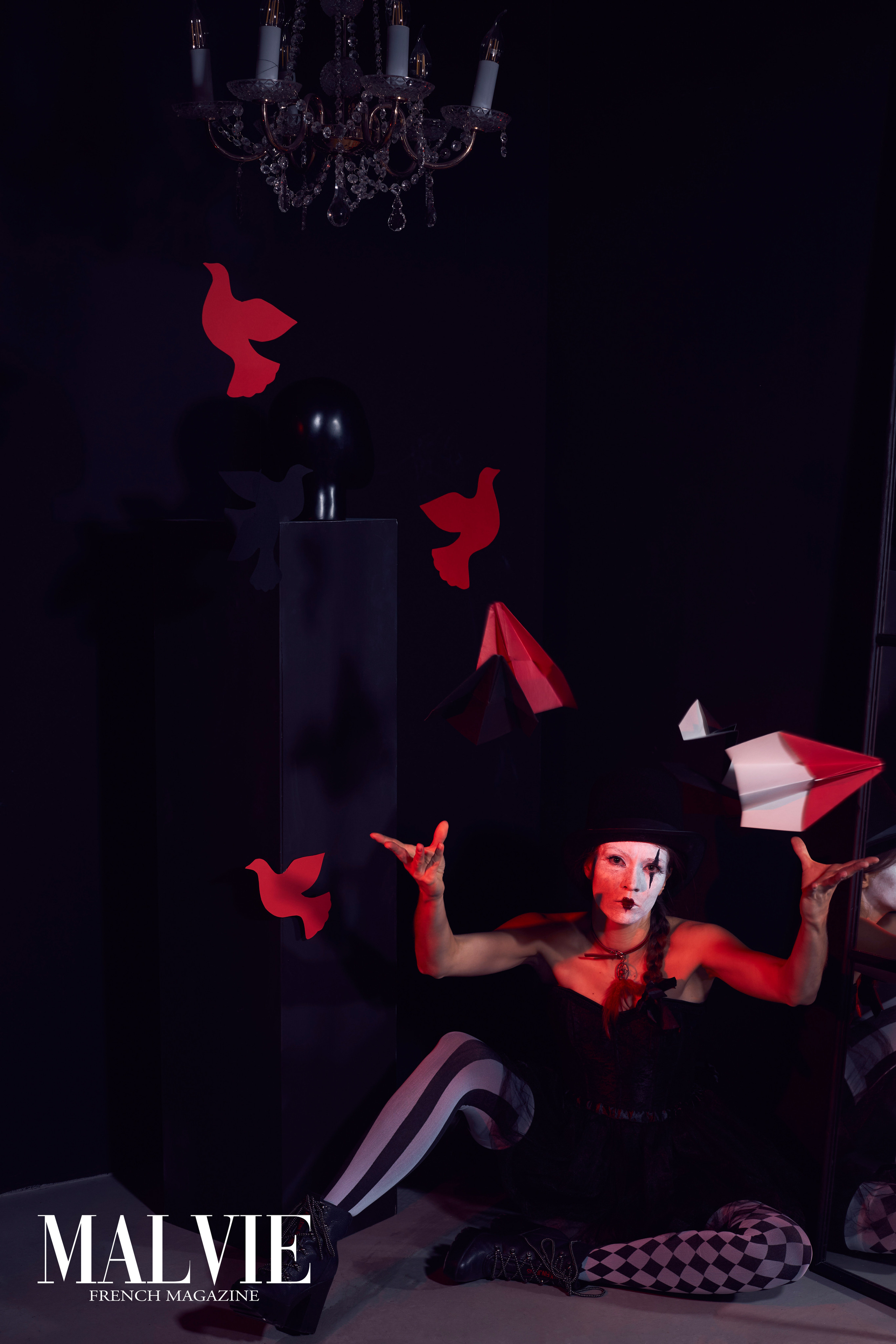 Девушка в черно-белом костюме арлекины или клоунессы сидит на полу и запускает красные бумажные самолётики