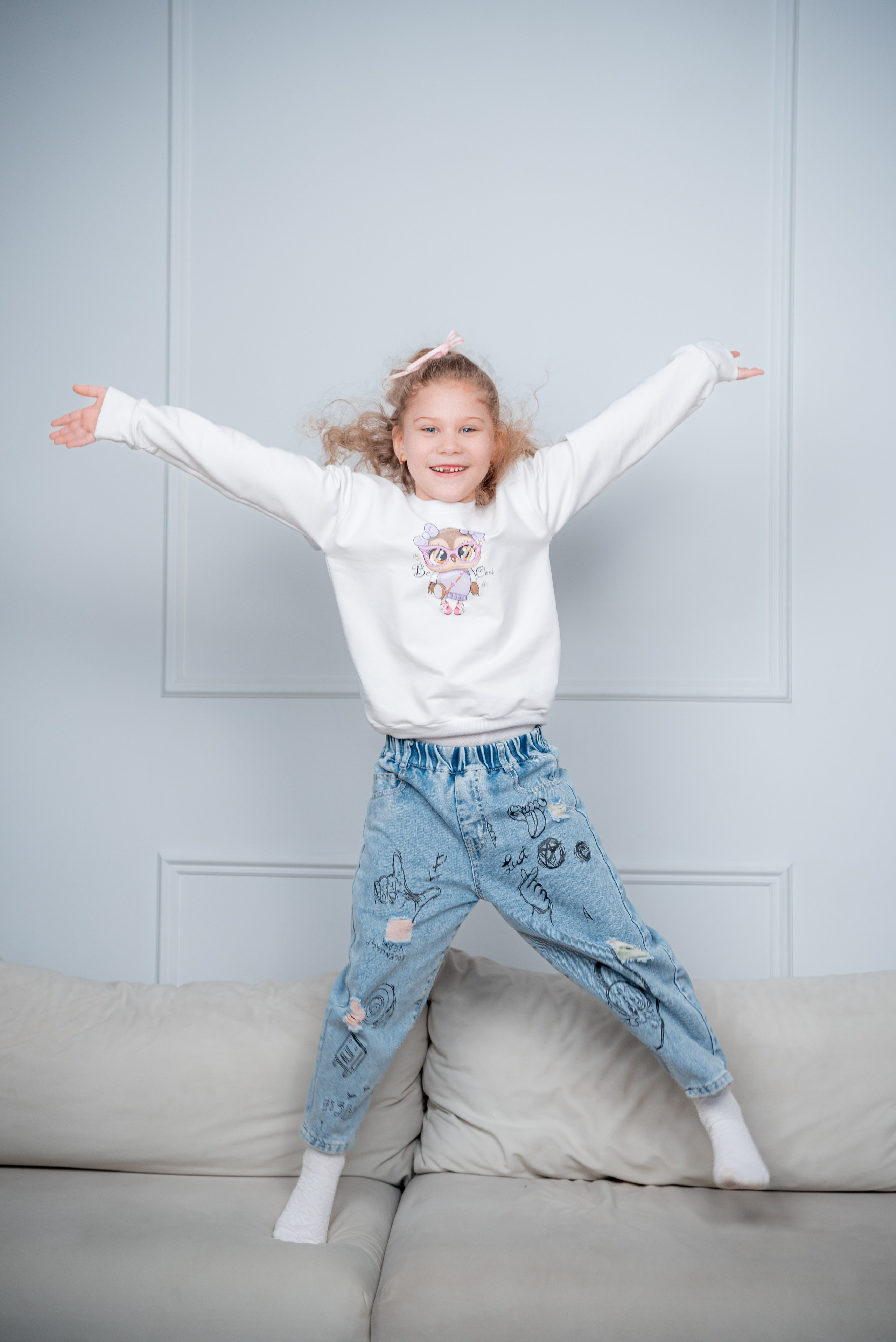 Младшая дочка прыгает на диване в белой кофте и синей джинсах
