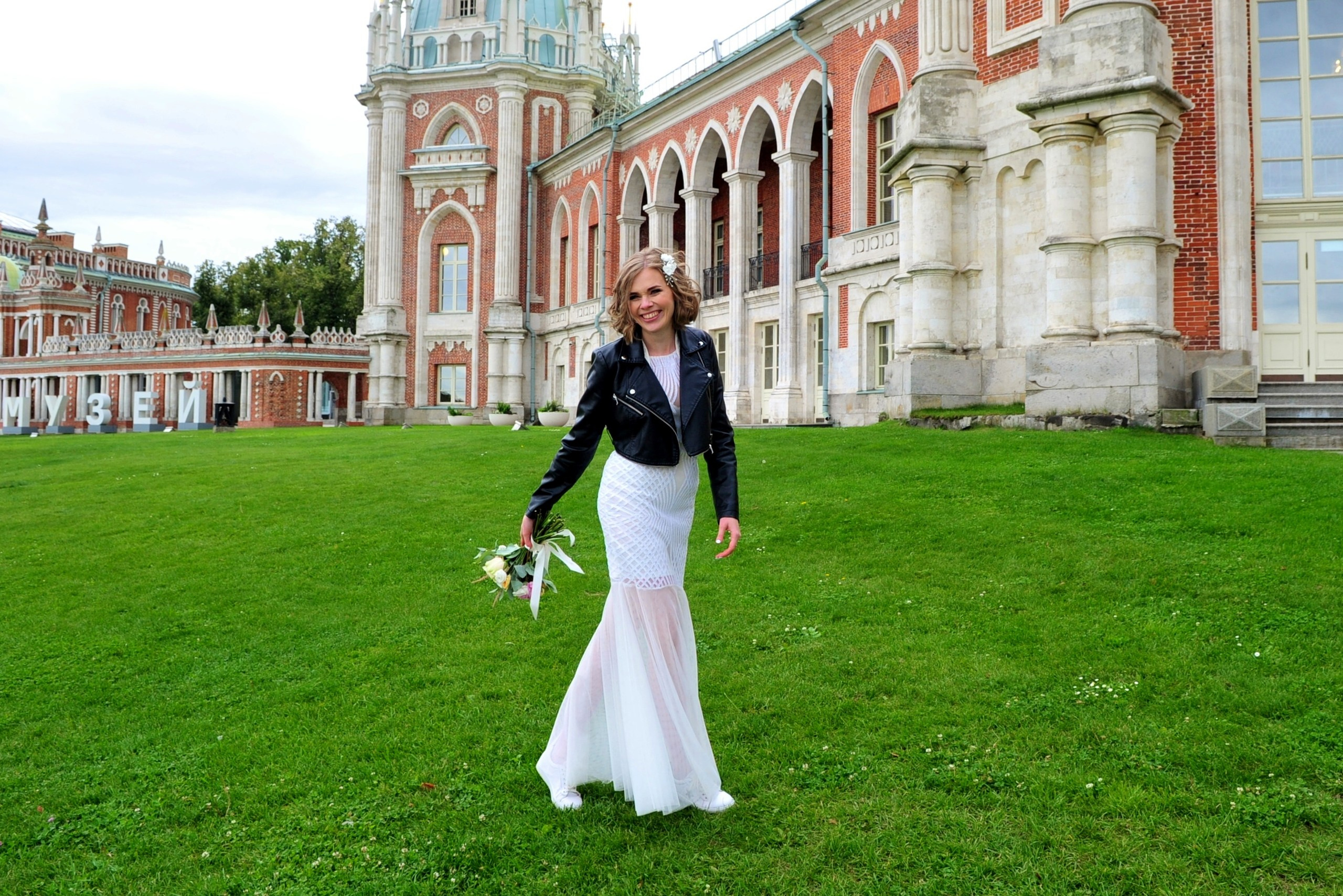 Свадьба в Царицыно хмурым прохладным летним днём, довольная невеста идет по зеленой лужайке на фоне дворца из красного кирпича и белого камня в парке