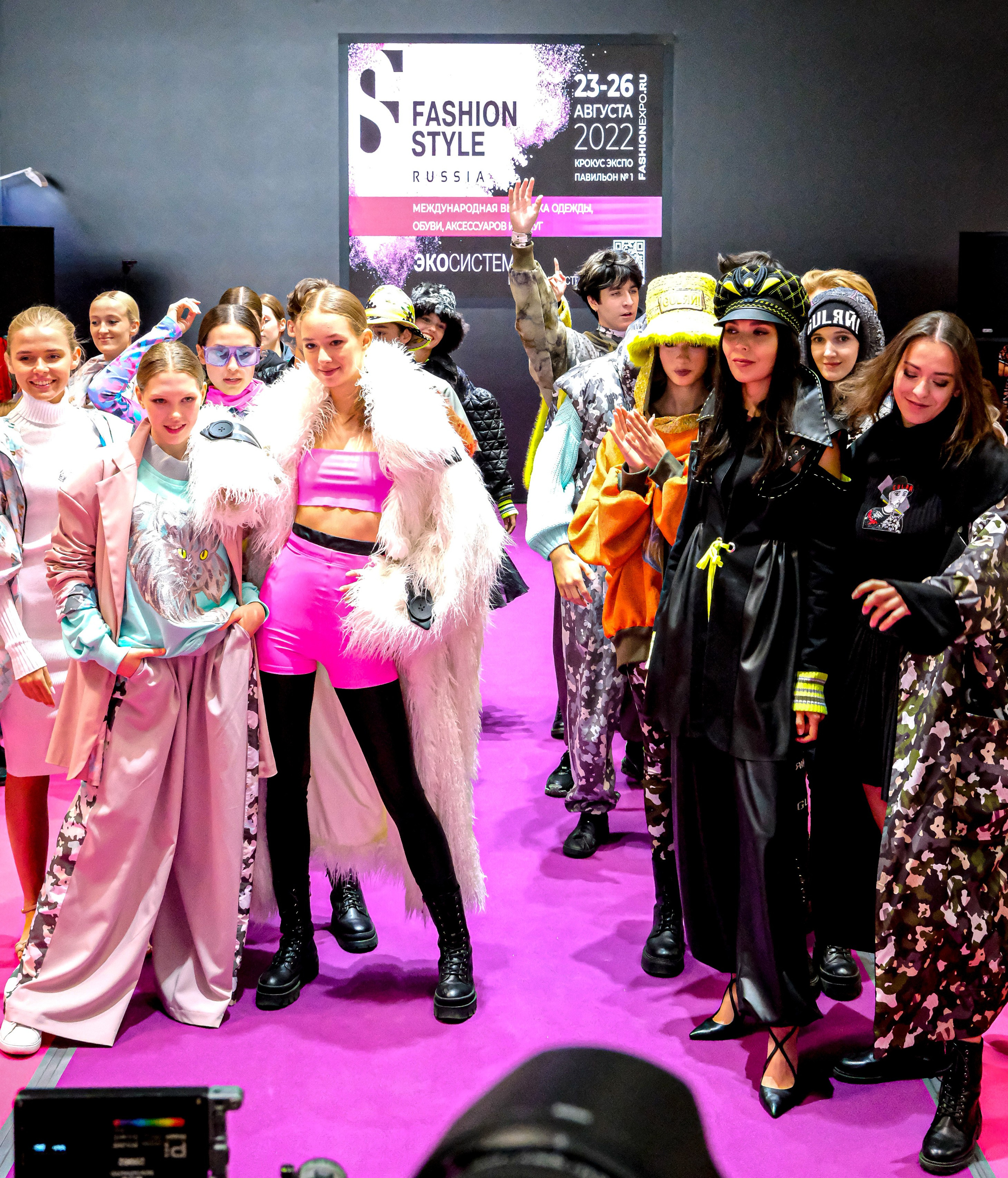  "Модели в ярких нарядах проходят по подиуму во время показа мод, освещенные яркими софитами и окруженные восторженными зрителями."