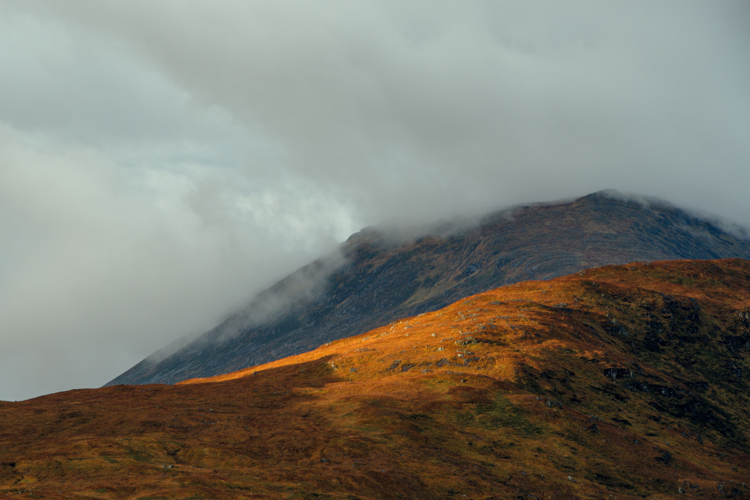 Драматический пейзаж острова Скай, Шотландия: пологий склон холма, освещенный золотым светом на переднем плане, и темные горы, окутанные низкими облаками на заднем плане.