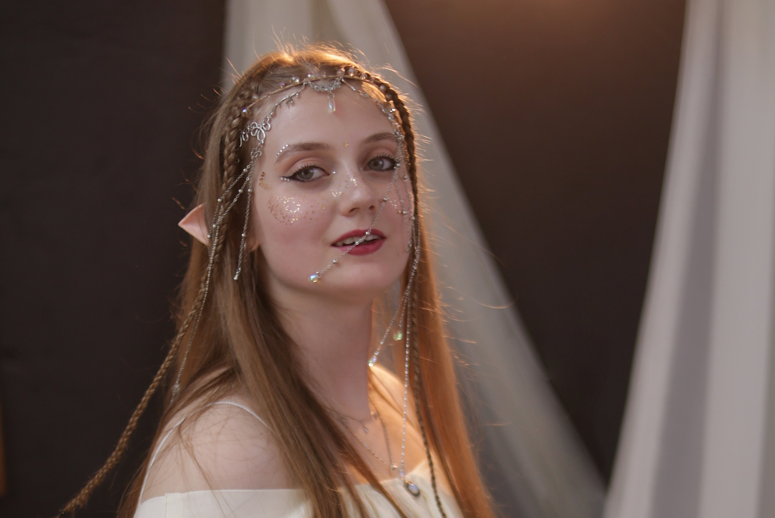 Крупный план эльфийки: девушка в белом платье с ушками и головным убором, лицо украшено блестками