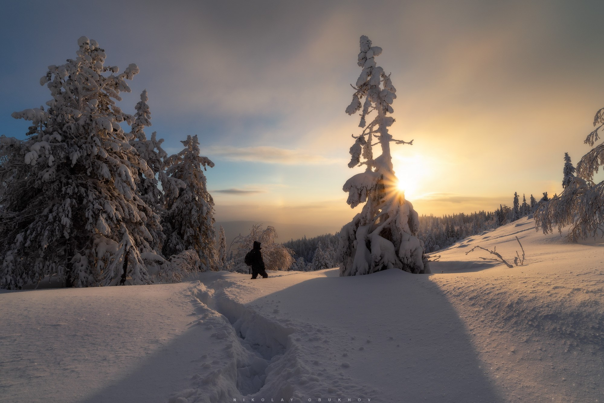 Фото зимнего пейзажа на горнолыжном комплексе «Гора Белая» в Свердловской области: восходящее солнце, человек на снежном склоне и  заснеженный лес.