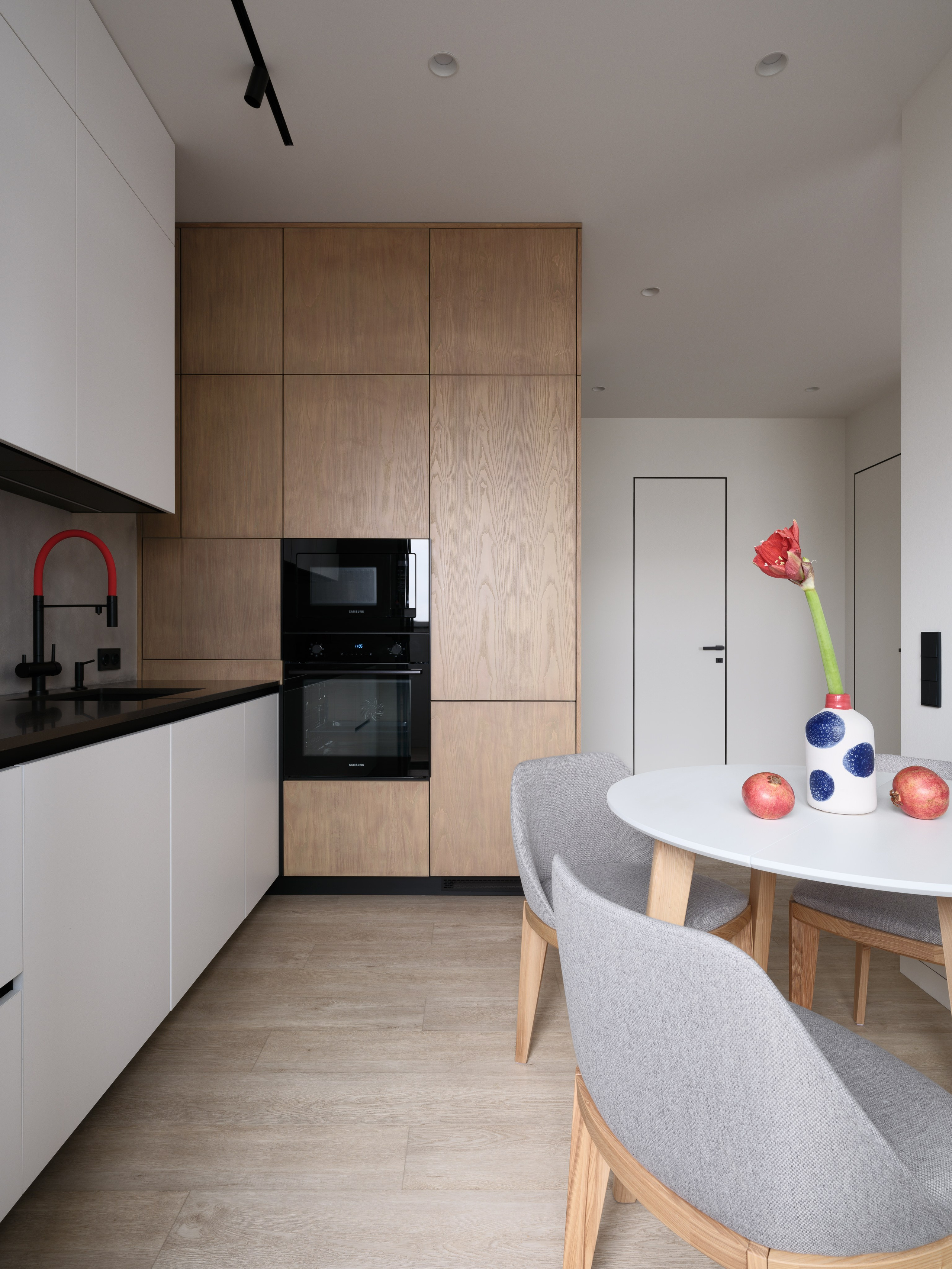 Современная кухня в минималистичном стиле с деревянными фасадами, встроенной техникой, белыми нижними шкафами и обеденной зоной с круглым столом и серыми стульями. Автор проекта — дизайнеры Евгения Княжева и Владлена Привалова, фотограф Сергей Денисенко
