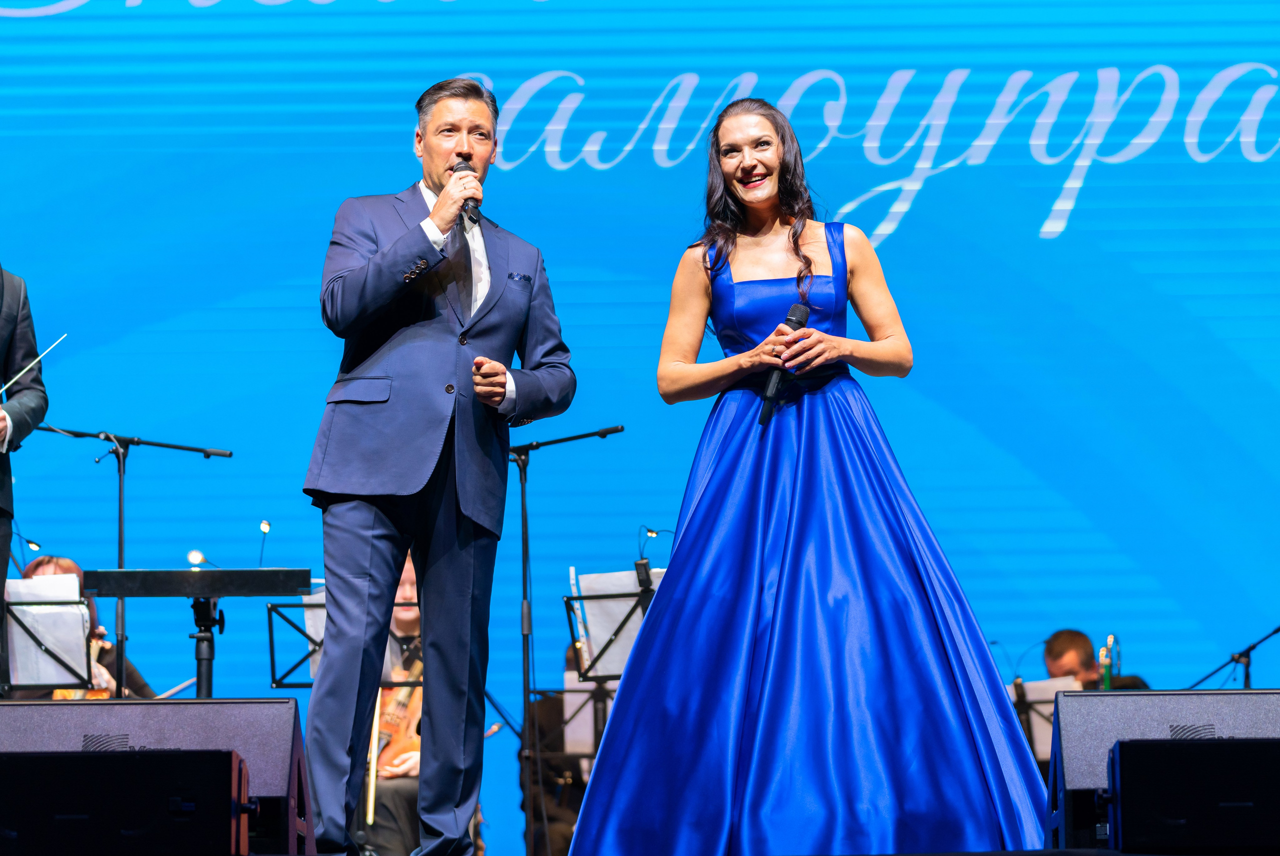 Ведущая  в синем платье и ведущий в синем костюме выступают дуэтом на сцене. Концерт ко Дню местного самоуправления Санкт-Петербурга, 2026. Фото Евгения Мальцева.