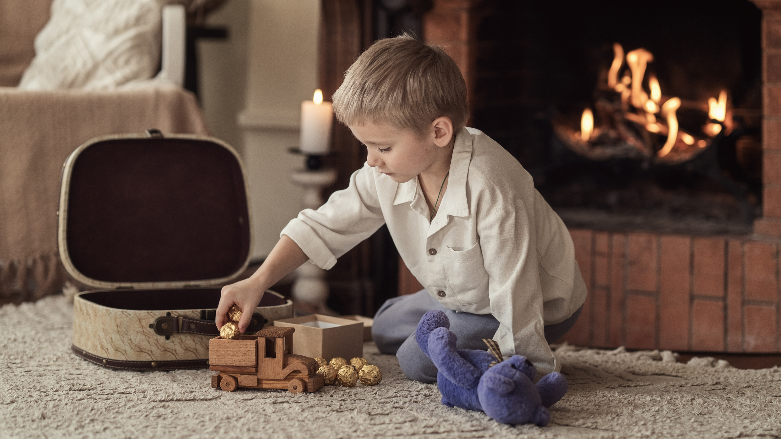 Мальчик у камина играет с плюшевым мишкой и деревянной машинкой, грузит в нее шоколадные конфеты