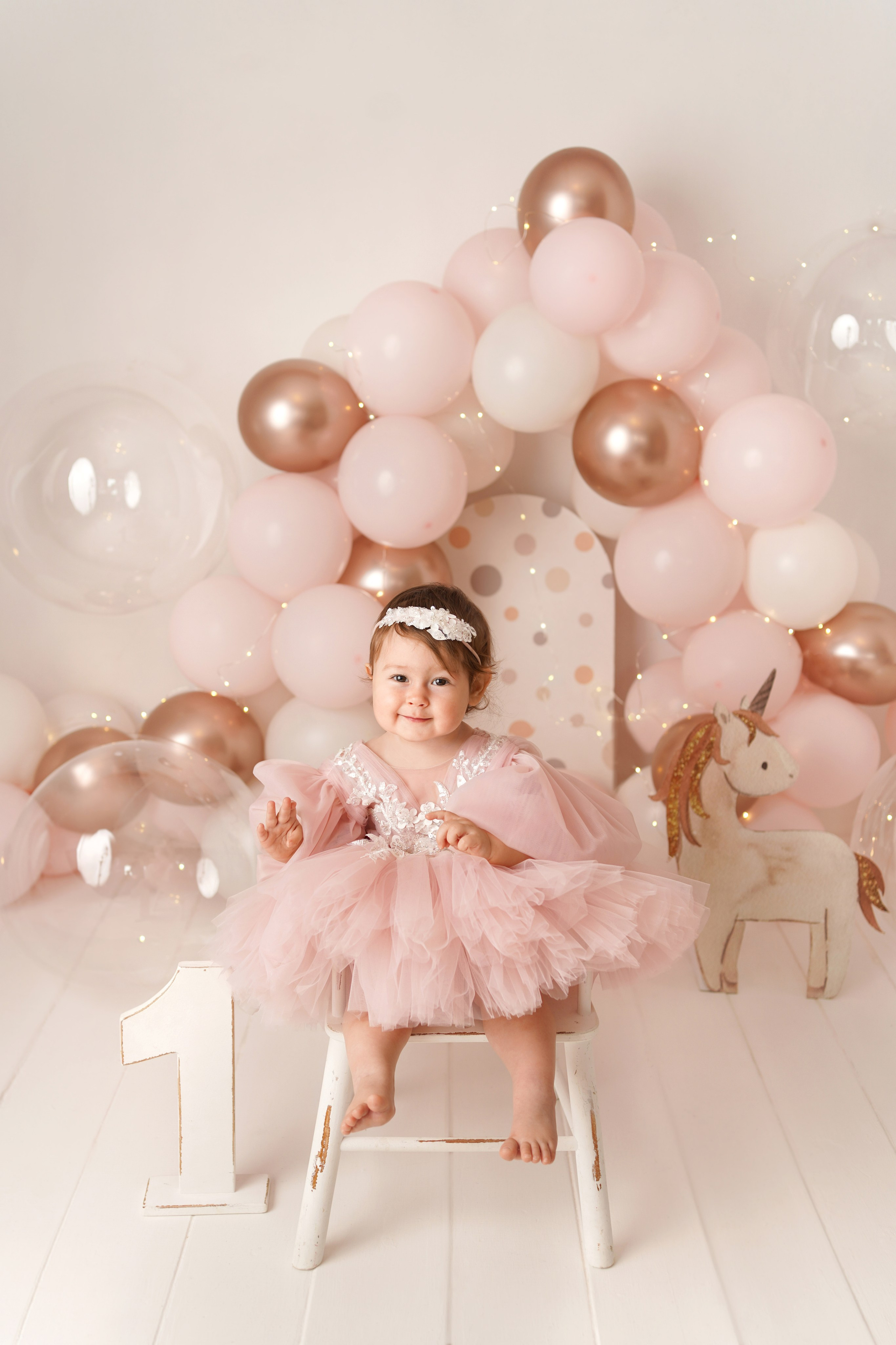 На фото малышка сидит на стульчике в праздничной фотозоне с шариками.