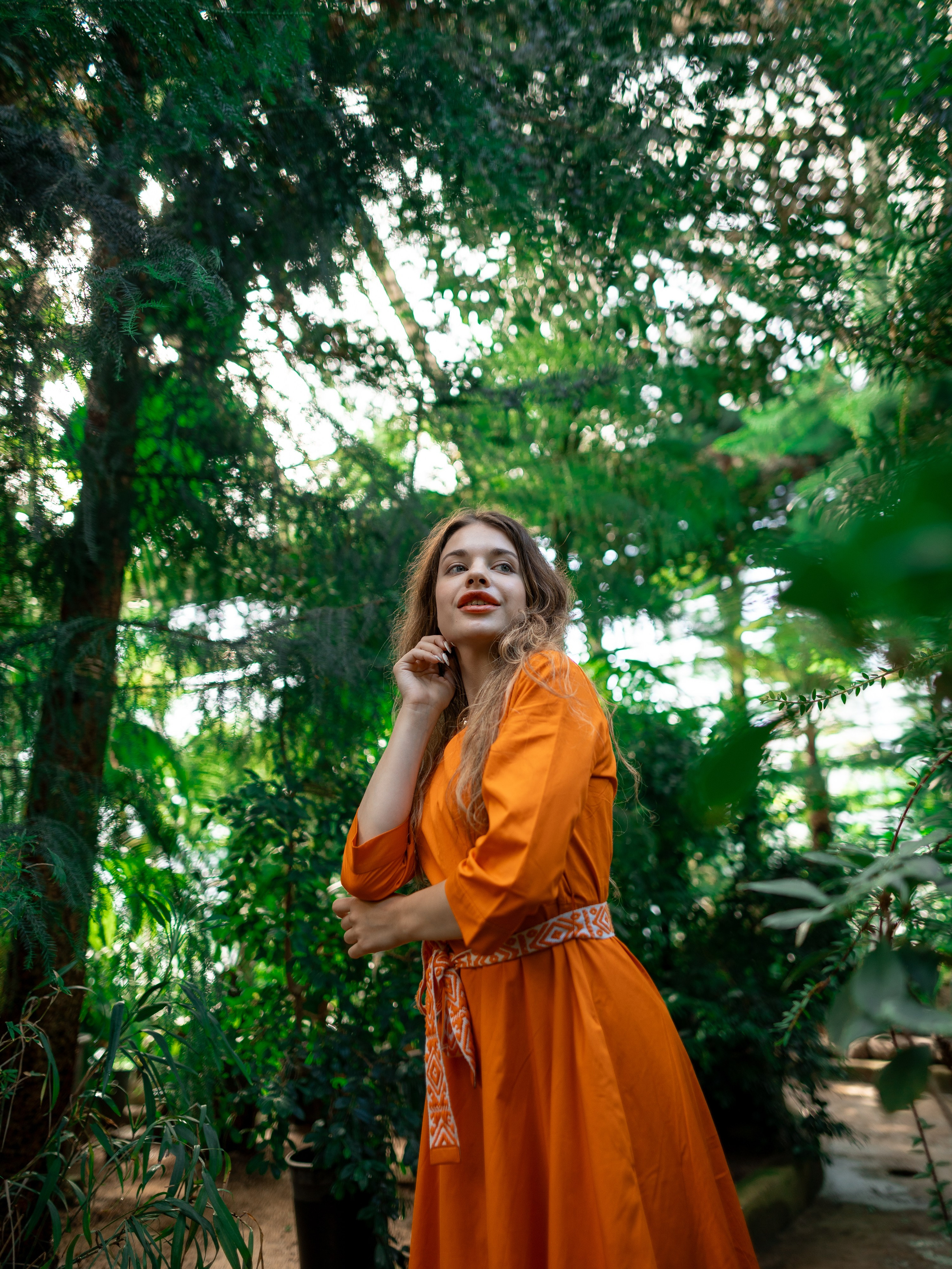 Девушка в оранжевом платье стоит на фоне зелени