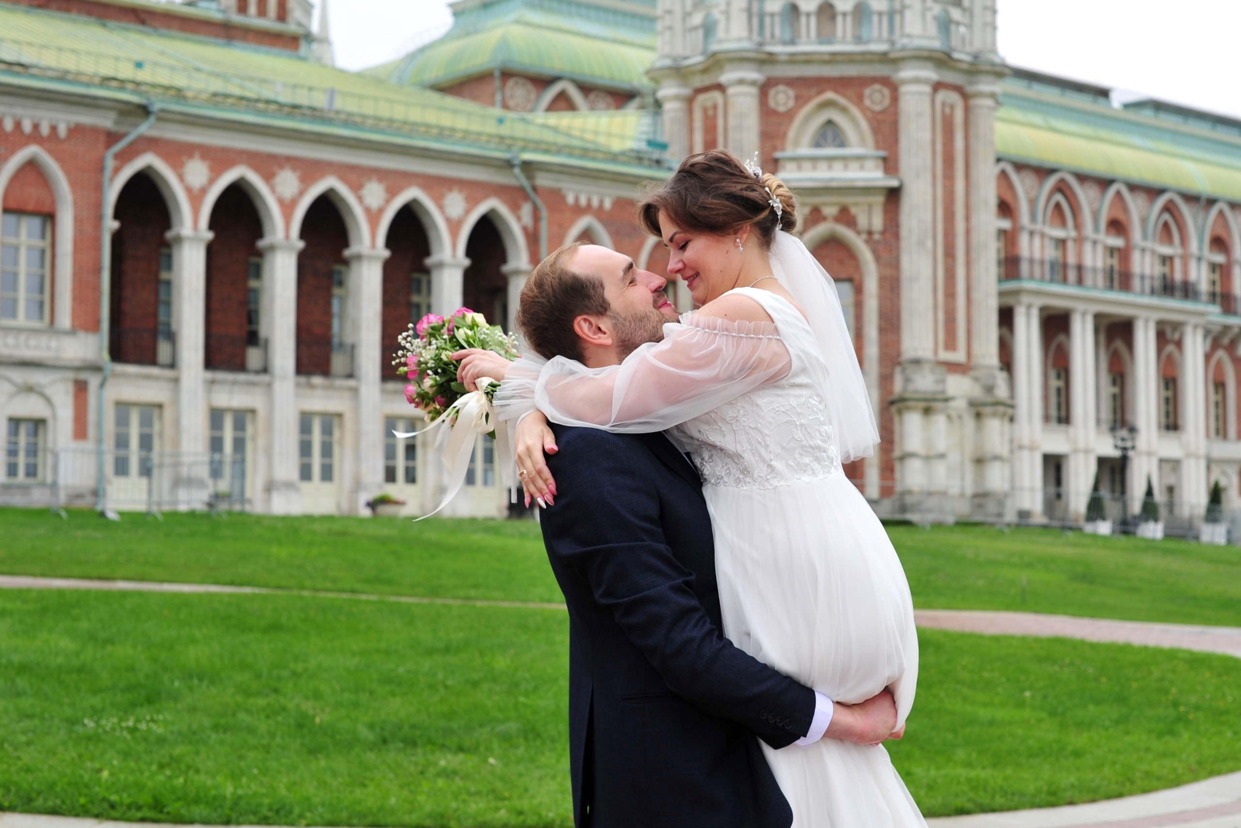 Свадьба в Царицыно пасмурным летним днем, жених держит невесту на руках на фоне дворца из красного кирпича и белого камня 