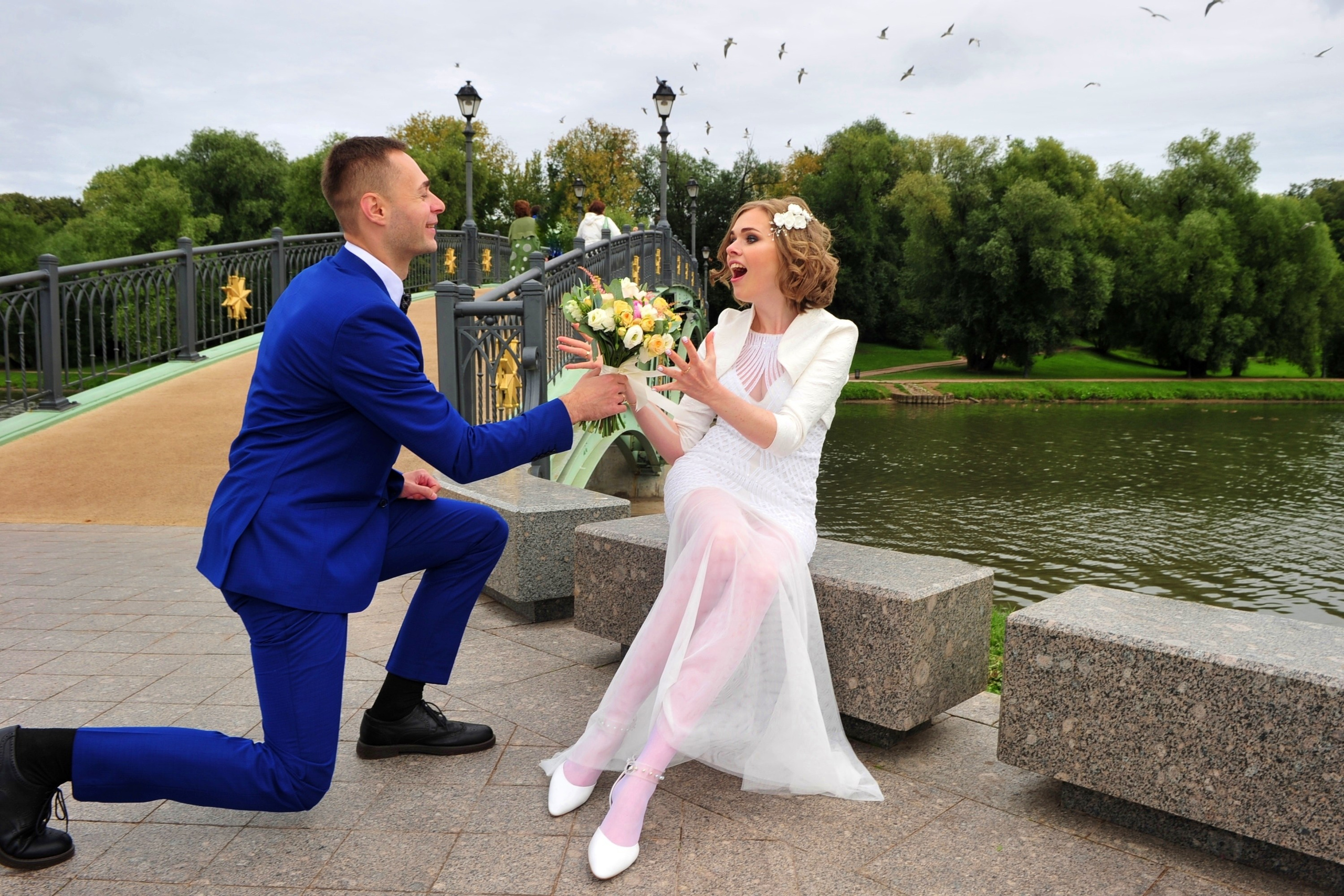 Свадьба в Царицыно хмурым прохладным летним днём, жених стоит на одном колене перед невестой и дарит ей букет, невеста удивлена и смеётся, молодожены на фоне пруда и железного моста