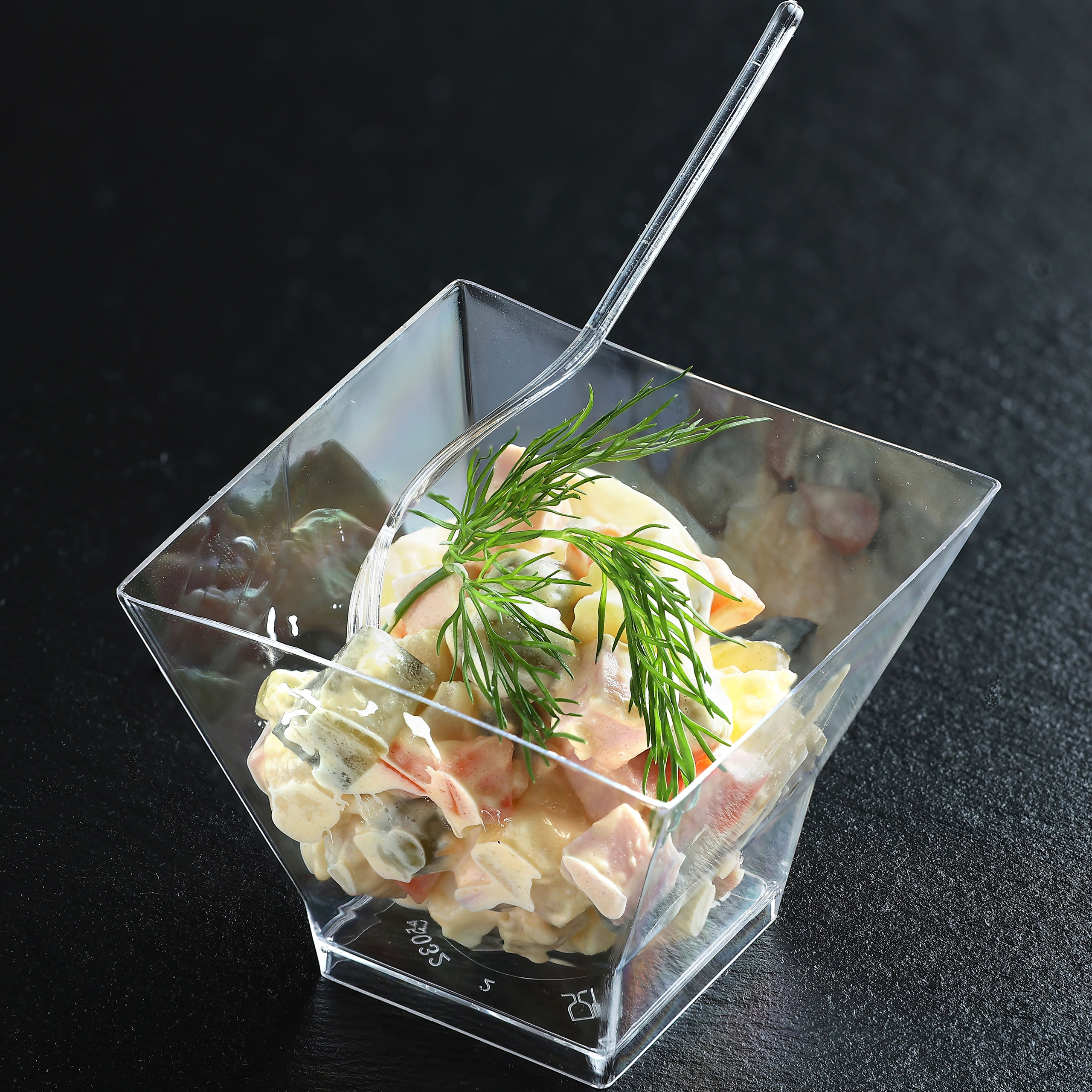 Классический салат Оливье с докторской колбасой и укропом в одноразовой пиалке с вилкой.