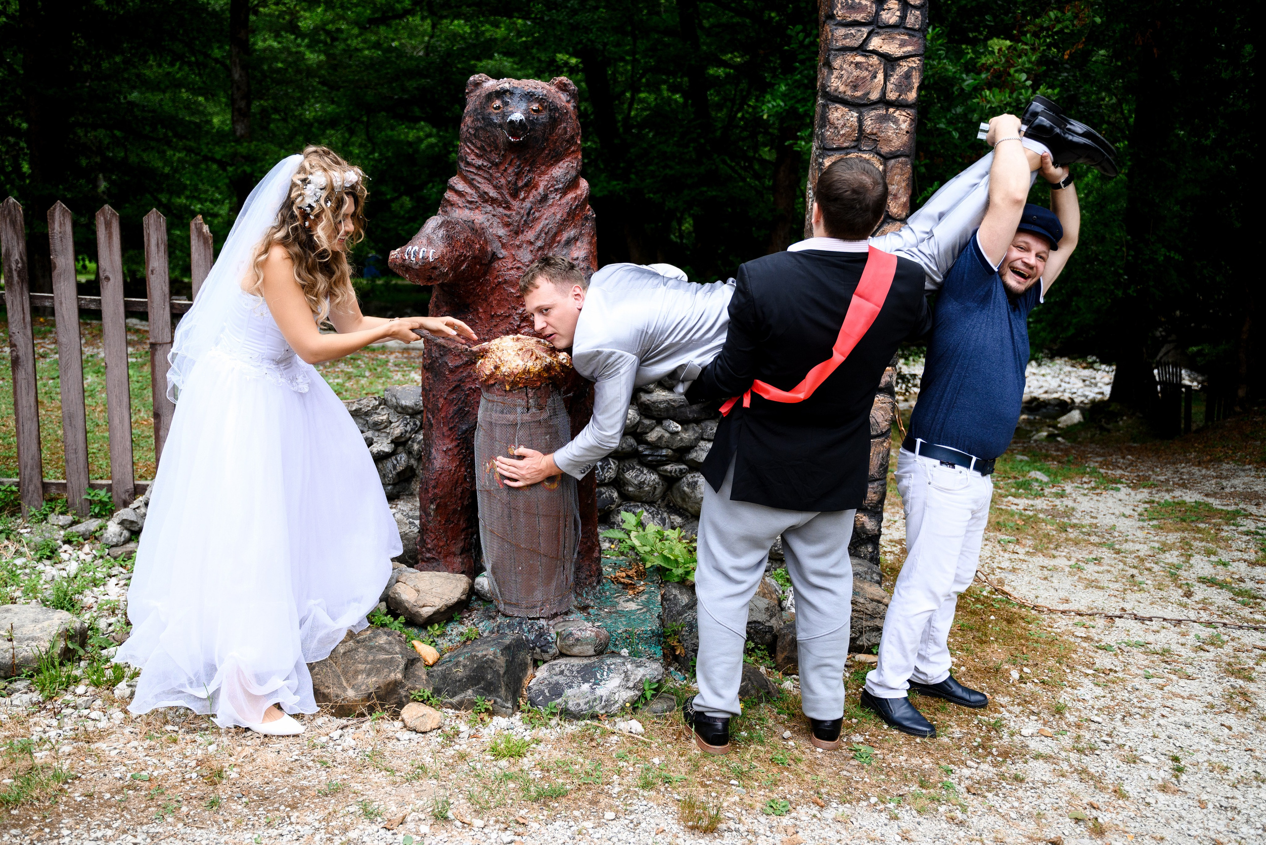 двое гостей взяли жениха на руки слева стоит невеста у какого-то памятника вырезанного из дерева форме медведя все это происходит в лесу 