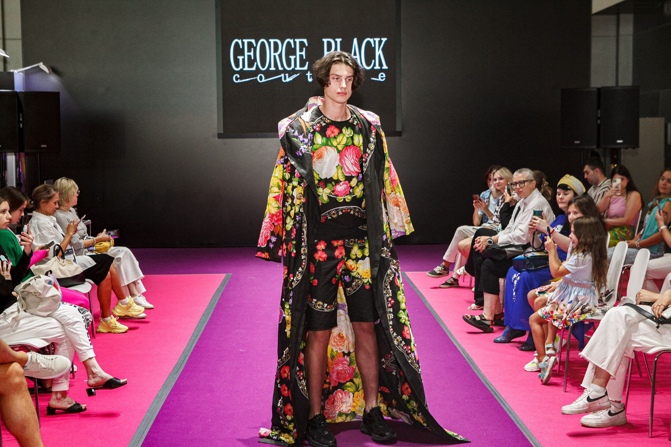  "Модели в ярких нарядах проходят по подиуму во время показа мод, освещенные яркими софитами и окруженные восторженными зрителями."