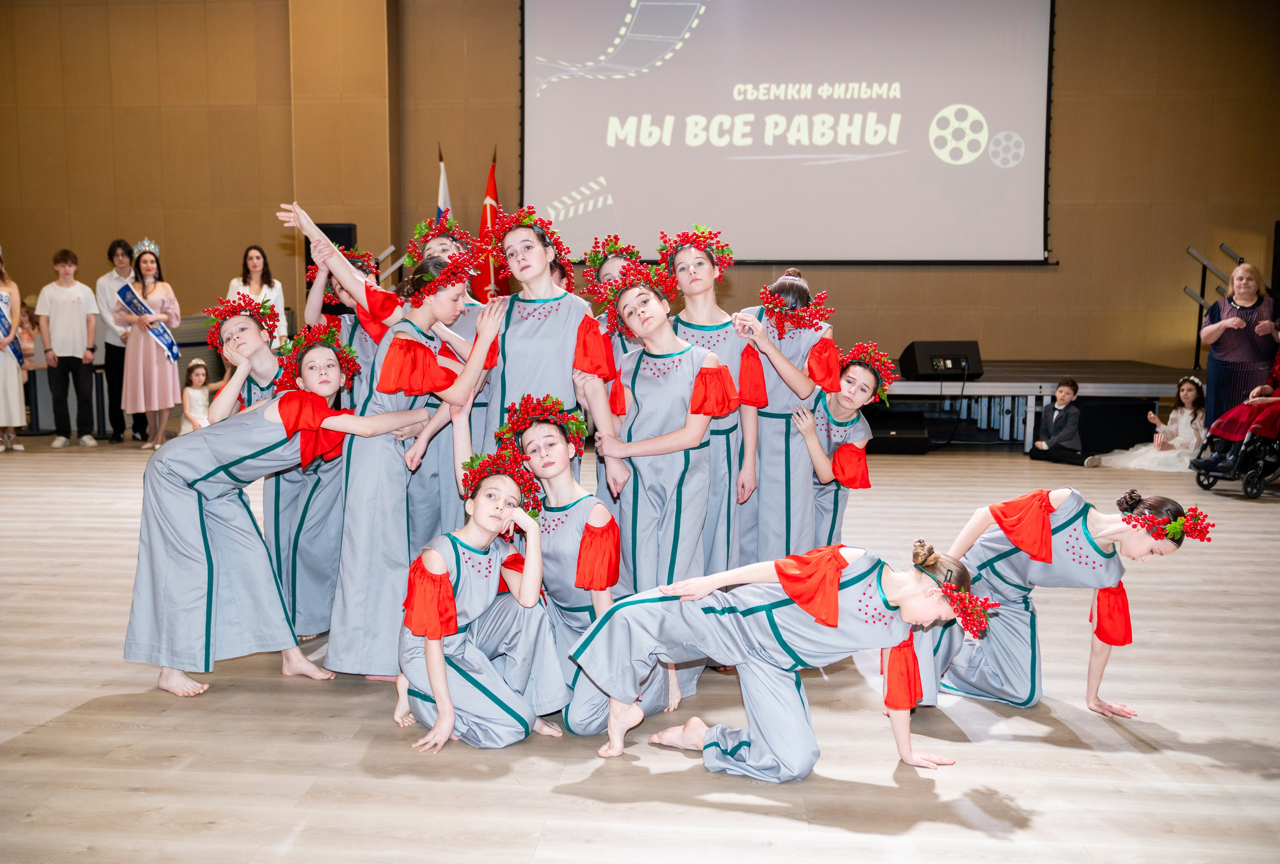 Группа девушек в серо-красных костюмах с венками из красных ягод исполняет танец на инклюзивном балу проекта «Мы все равны». Фотограф Мальцев Евгений.