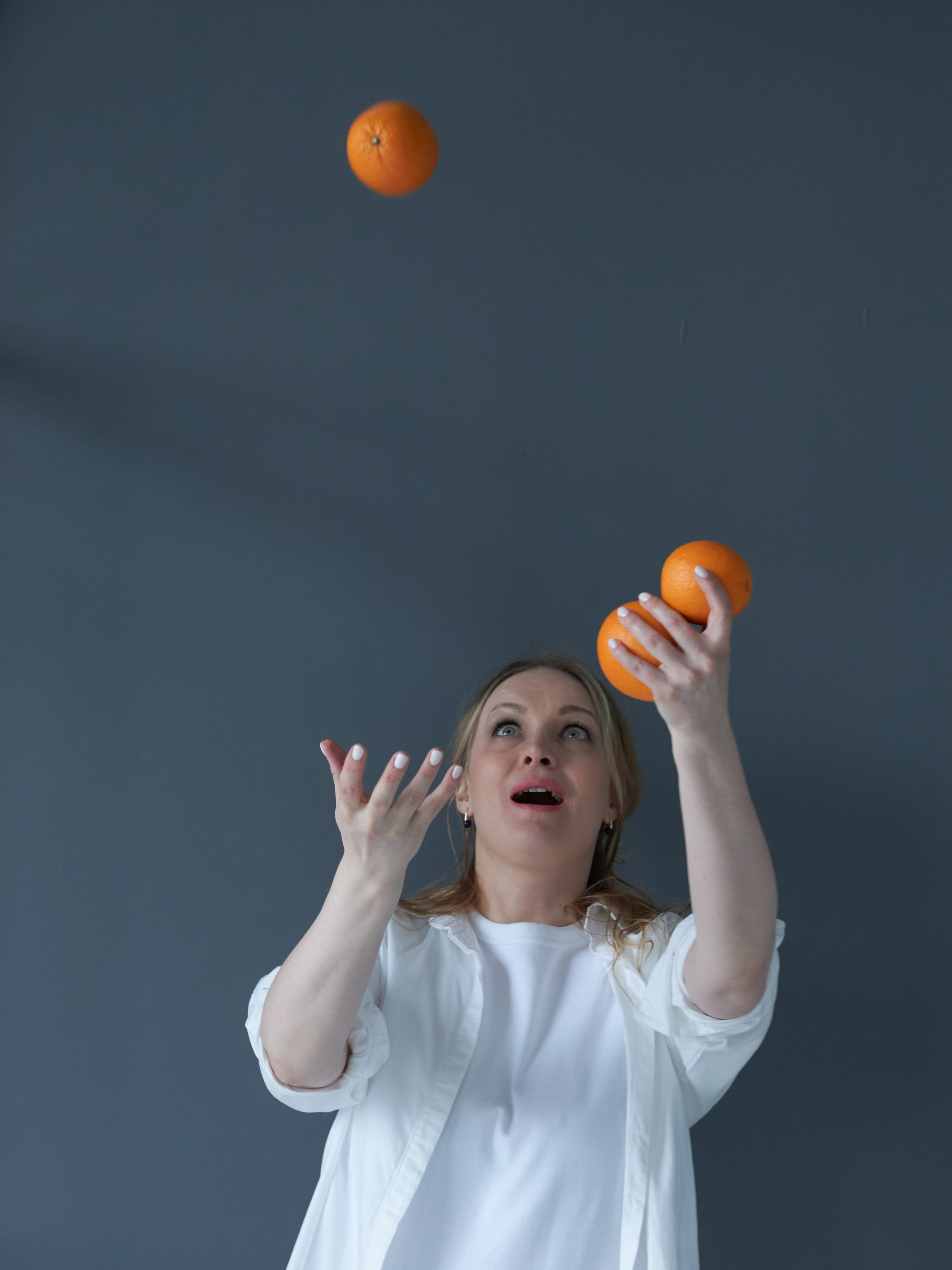 Девушка жонглирует апельсинами на фоне серой стены
