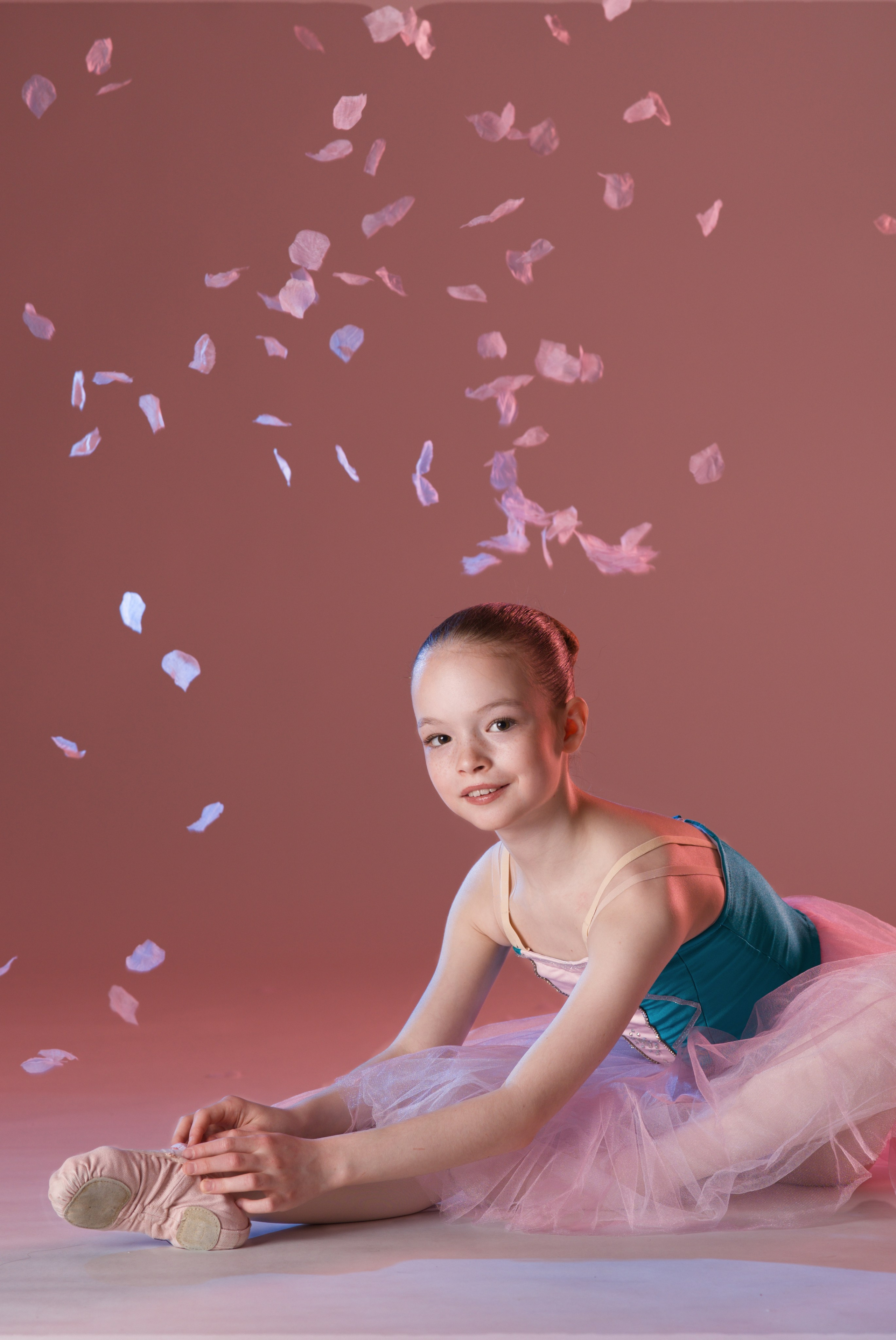 Юная балерина сидит на продольном шпагате в окружении летящих лепестков цветов