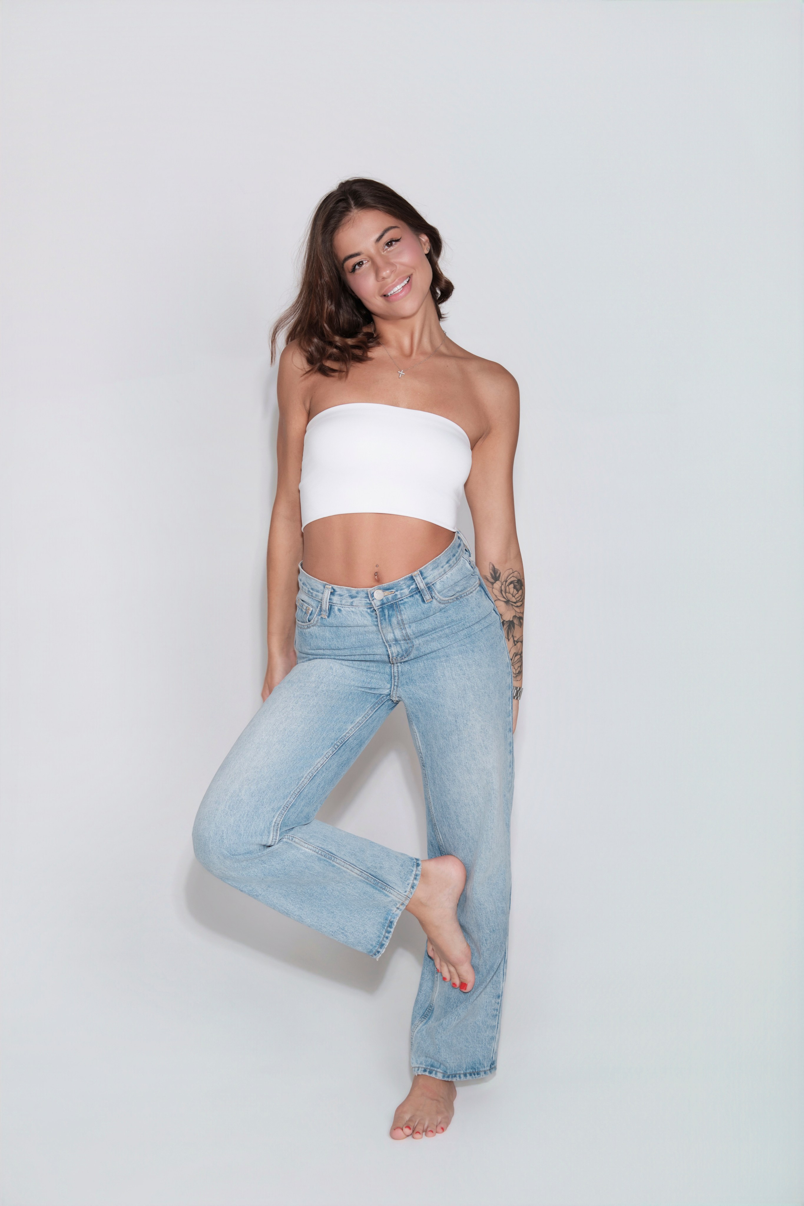 Студийная съемка модели в полный рост. Девушка позирует босиком в голубых джинсах и белом топе.